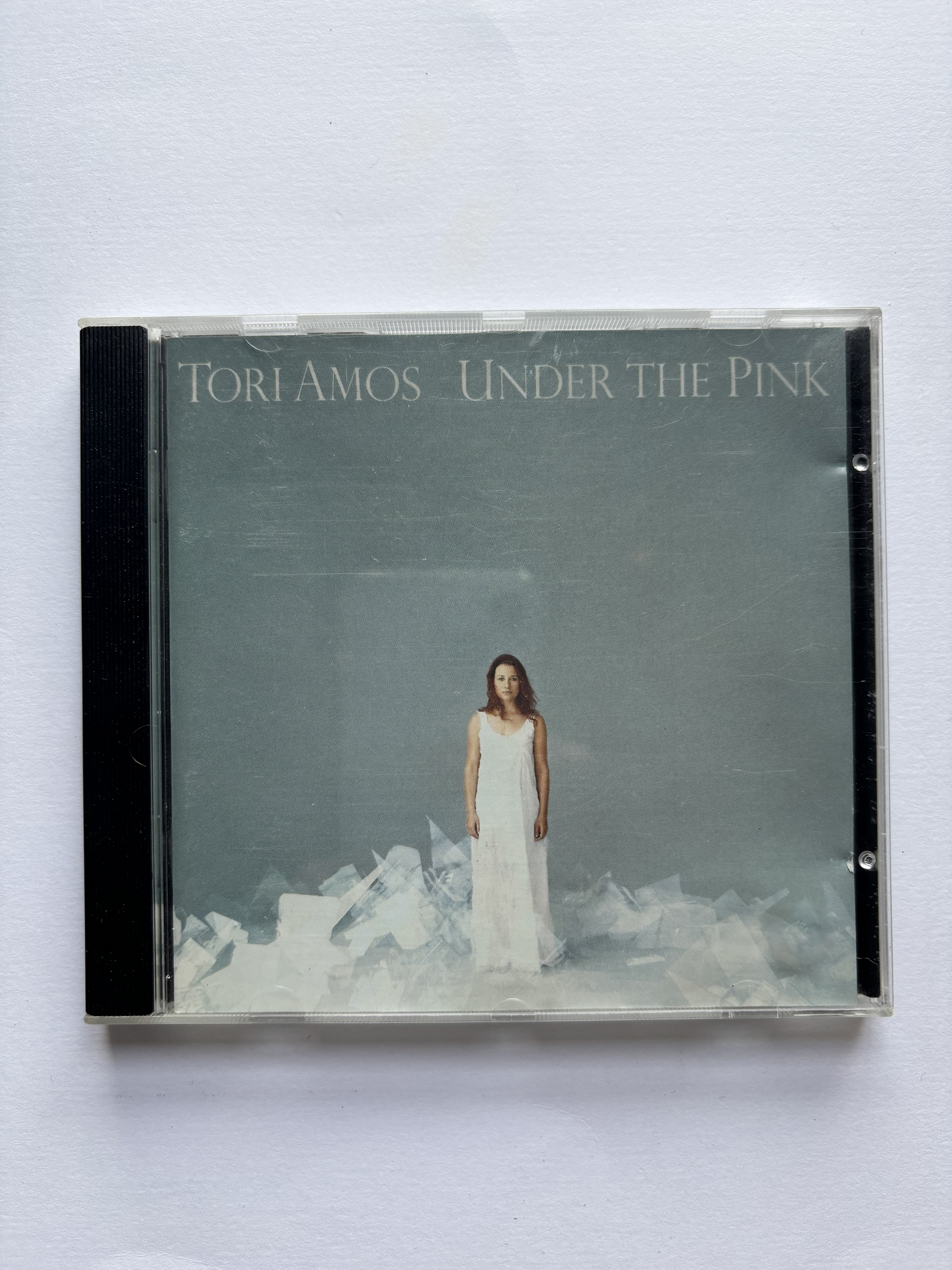 CD case for Tori Amos album titled 'Under the Pink' featuring a woman in a long white dress standing against a muted blue-gray background with scattered translucent shapes at her feet.