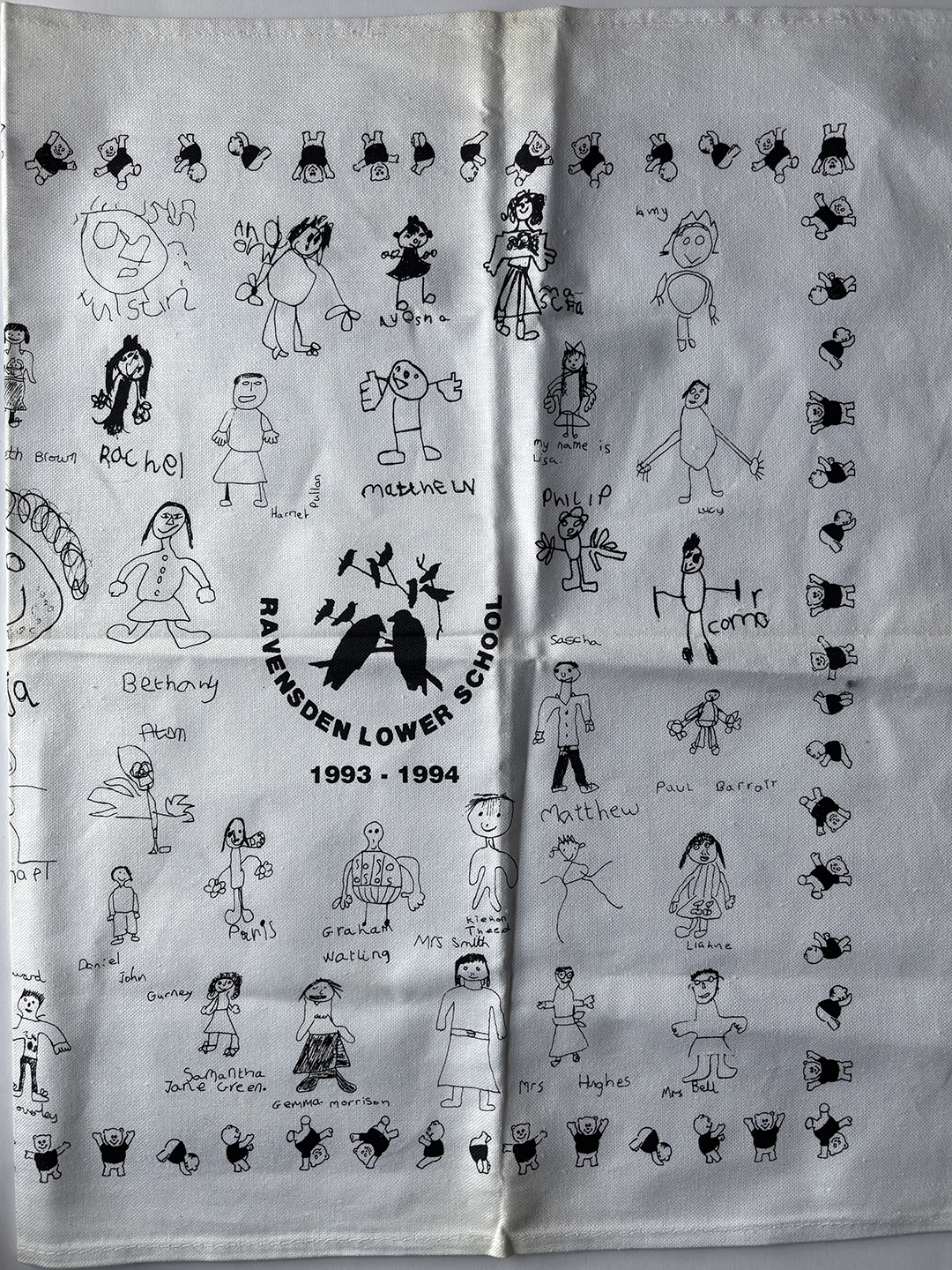 White tea towel printed with black childlike drawings of people and names, surrounding a central logo of birds on branches with text 'Ravensden Lower School 1993 - 1994'. Drawings are arranged around the edges and center, representing students and teachers.