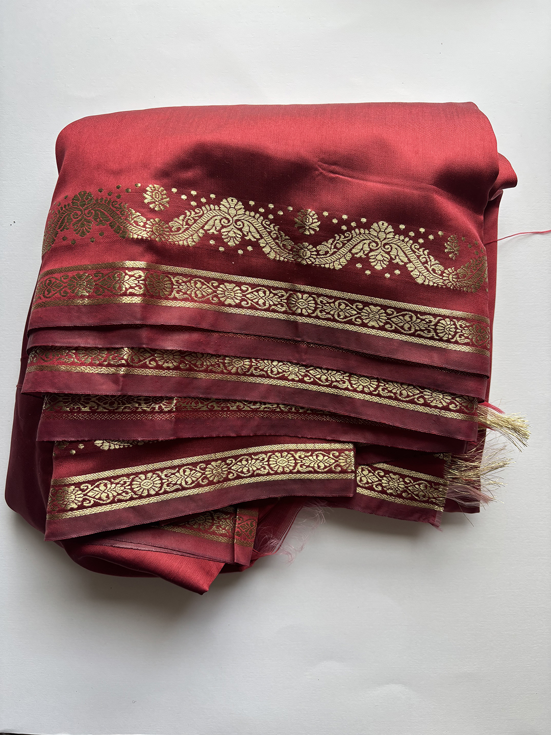 Folded deep red sari fabric with intricate gold floral and geometric border patterns laid on a white surface.
