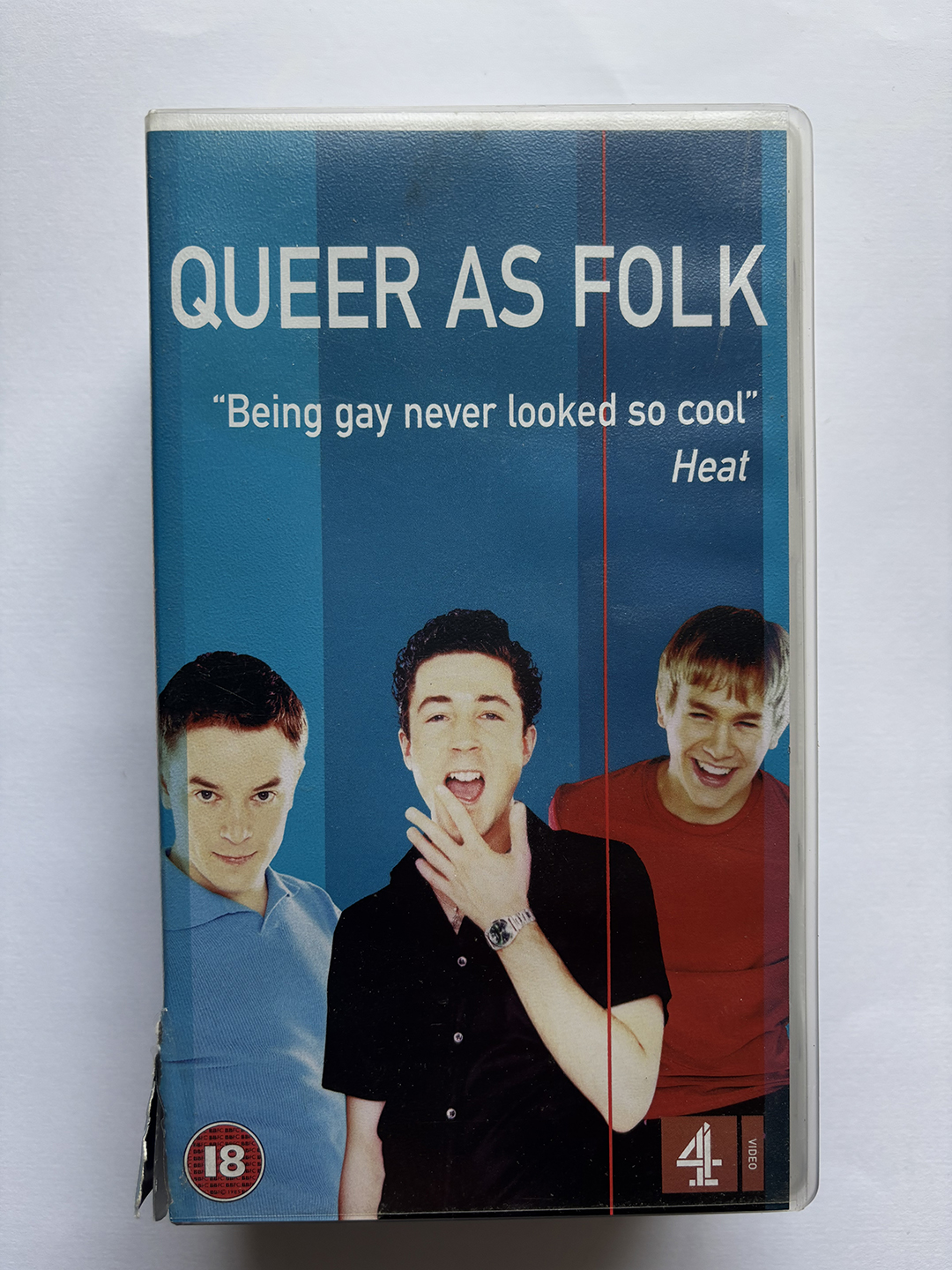 VHS cover of the TV show Queer As Folk featuring three young men against a blue striped background, with the quote "Being gay never looked so cool" attributed to Heat, an 18 age rating symbol, and the Channel 4 logo.