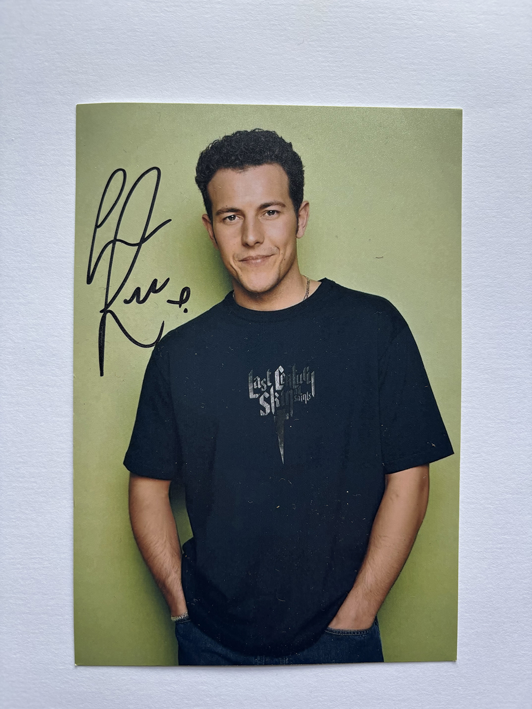 Young man with short curly hair wearing a black T-shirt with 'Last Sunday Saints' text, standing against a plain light green background with hands in pockets, smiling slightly, with a black autograph on the left side.