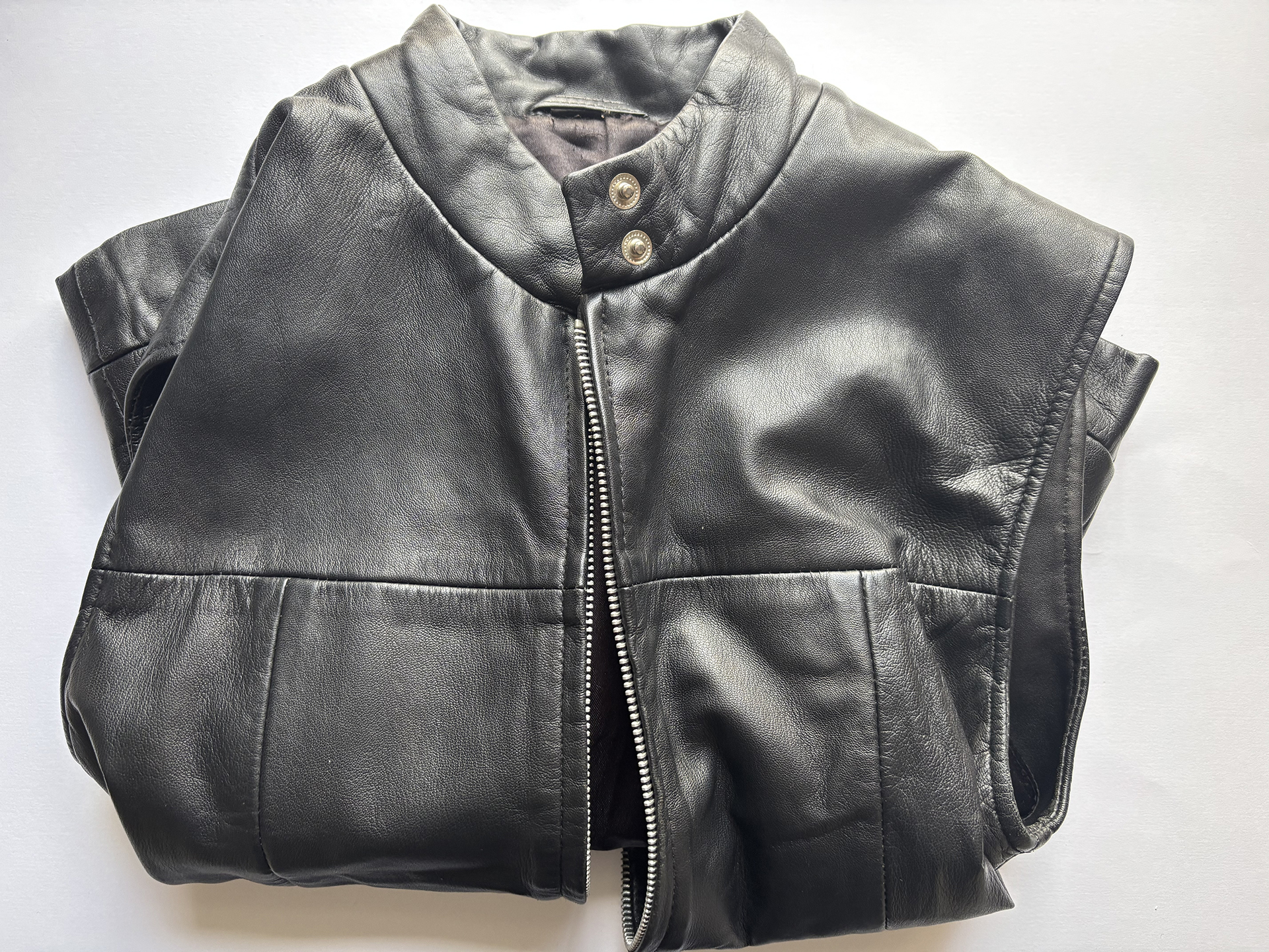 Folded black leather sleeveless waistcoat with a front zipper and two metallic snap buttons on the collar, displayed on a white background.