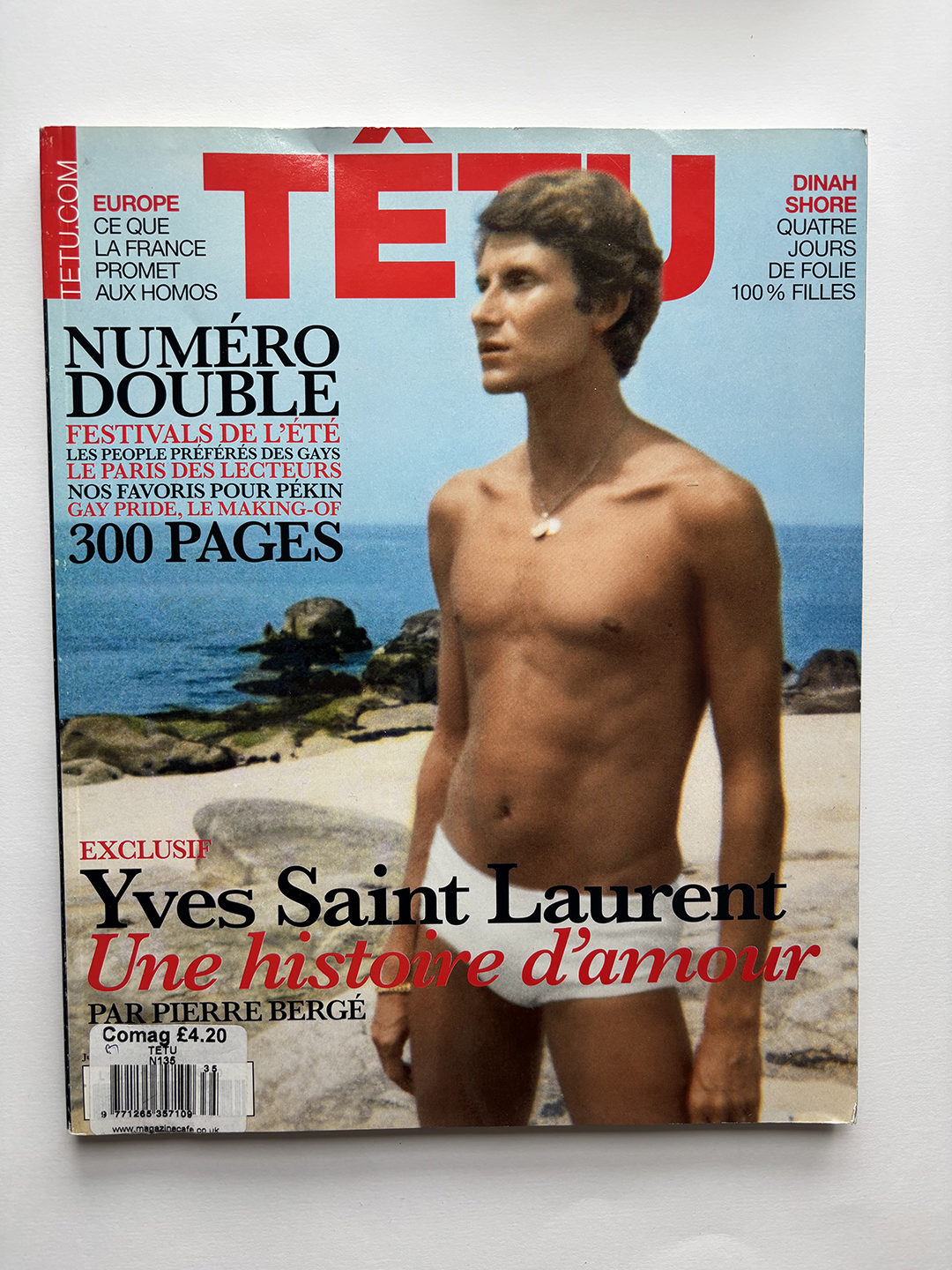 Cover of TÊTU magazine featuring a shirtless young man in white swim trunks standing on a rocky beach with the sea behind him, with text highlighting an exclusive story about Yves Saint Laurent and summer festivals.