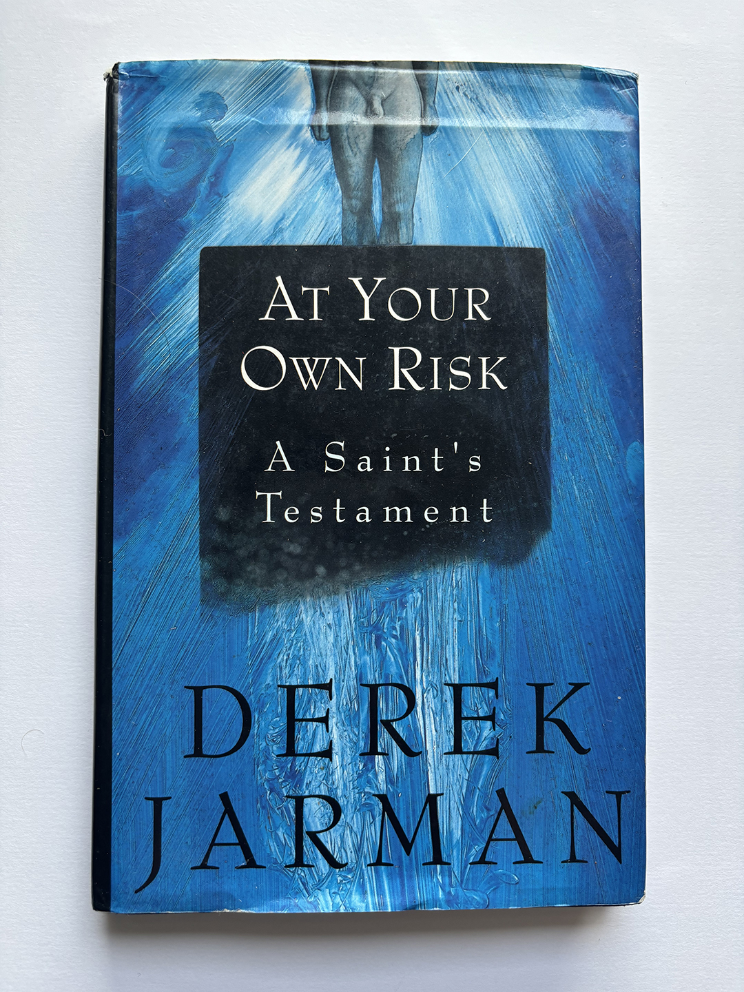 The cover of a book titled 'At Your Own Risk: A Saint's Testament' by Derek Jarman, featuring a blue background with a faint image of a standing, headless figure at top center.