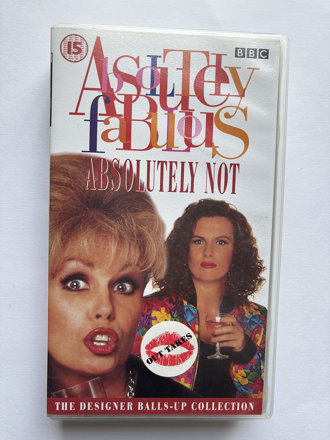 Cover of a VHS tape titled 'Absolutely Fabulous: Absolutely Not,' featuring two women; one with blonde hair and wide eyes holding a drink, the other with curly brown hair and a colorful jacket also holding a drink. The cover includes the BBC logo, a red sticker with red lips and 'OUT TAKES' text, and a red banner at the bottom that reads 'THE DESIGNER BALLS-UP COLLECTION.'