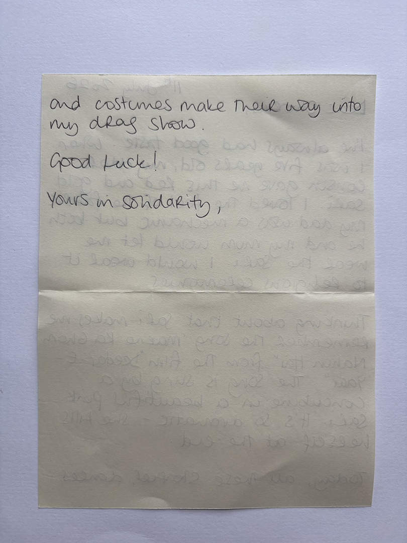 A handwritten note on a pale yellow paper, partially visible from the back side, reads: 'and costumes make their way into my drag show. Good luck! Yours in solidarity,'