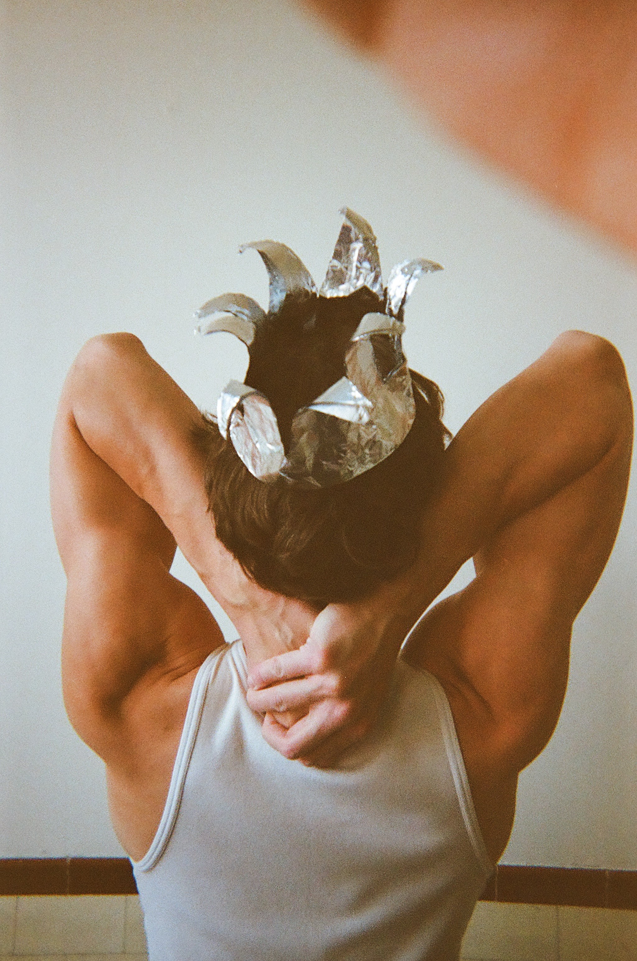 Person wearing a white tank top with muscular arms raised and hands clasped behind their head, wearing a silver foil crown on dark hair, shown from behind against a plain light background.