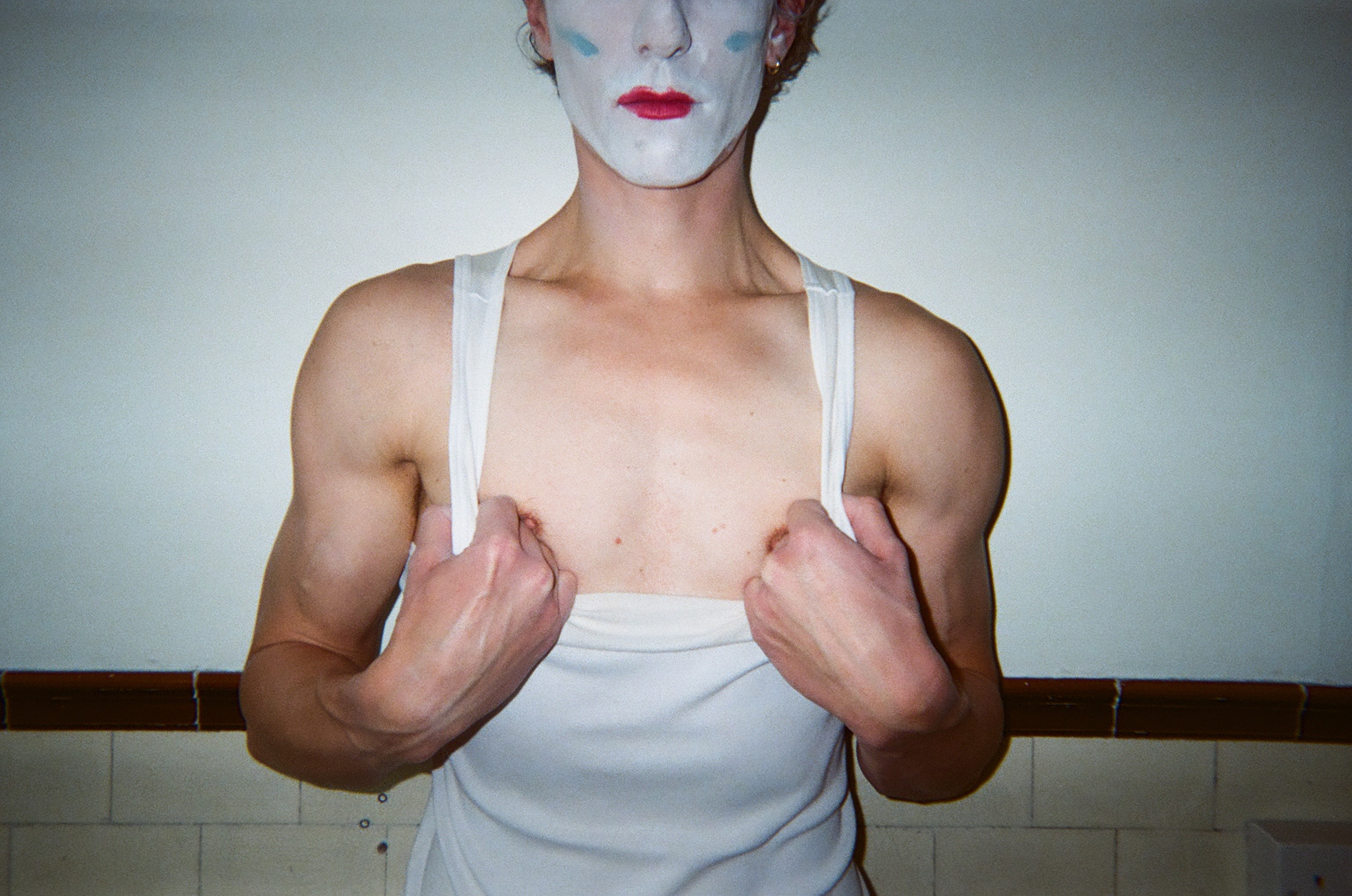 Person with theatrical white face paint, blue marks on cheeks, and red lipstick wearing a white sleeveless top that they are pulling down to reveal their chest and muscular arms.
