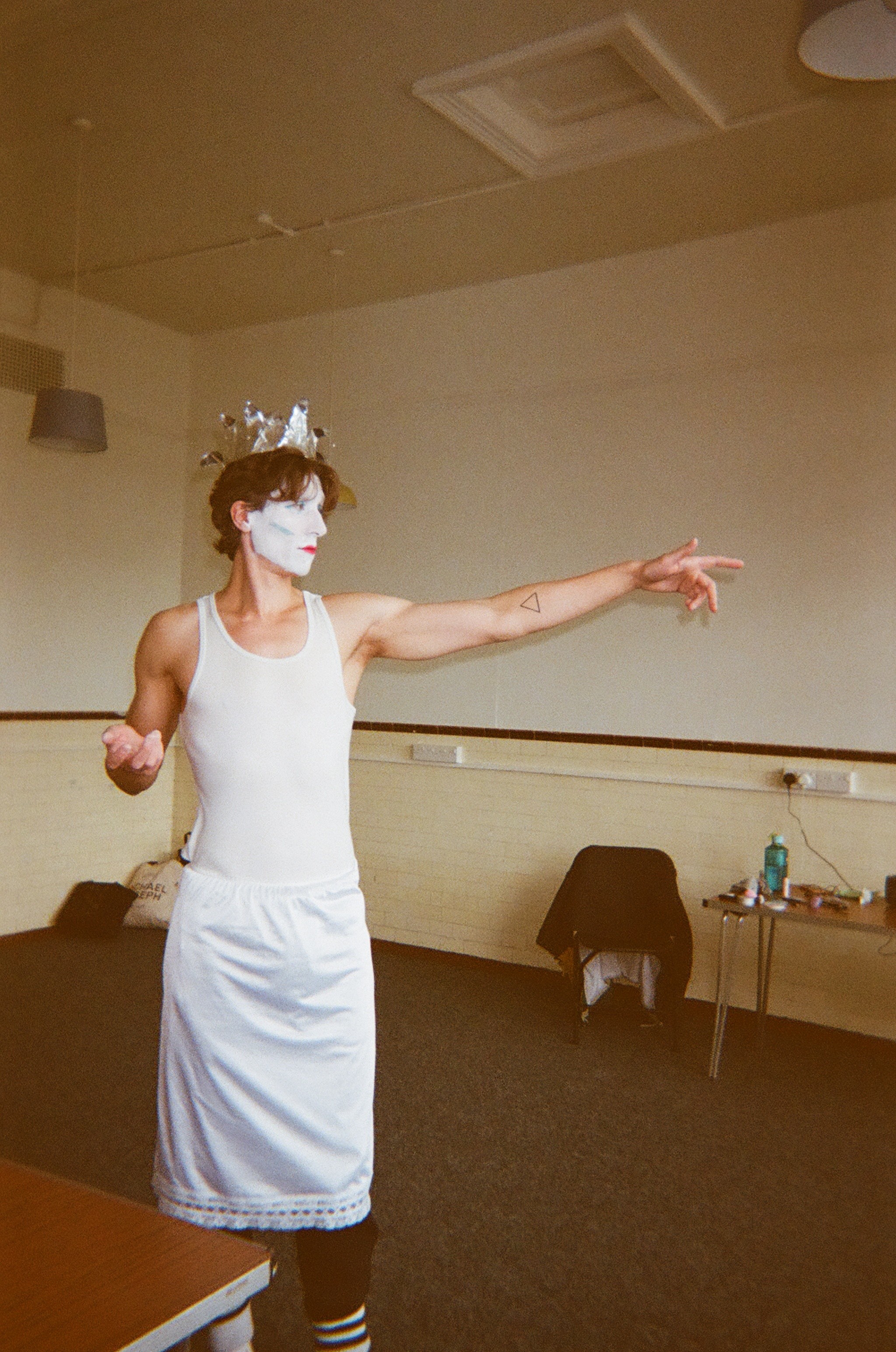 A person with white face paint and red lips wears a silver foil crown, a white tank top, and a white skirt, standing indoors with one arm extended and pointing.