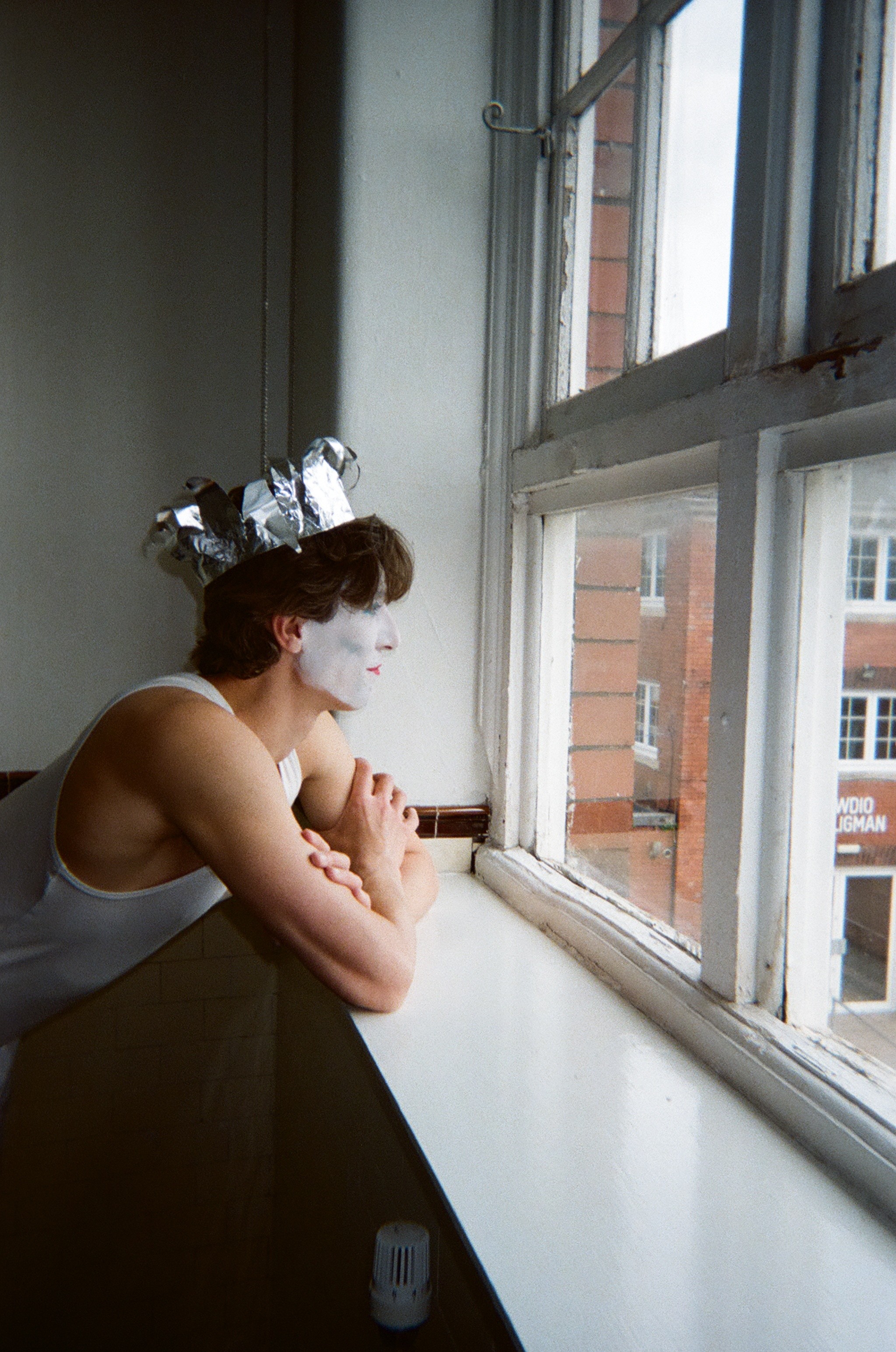 Person with white face paint and red lipstick wearing a silver foil crown, leaning on a windowsill and looking out a multi-pane window in a tiled room.