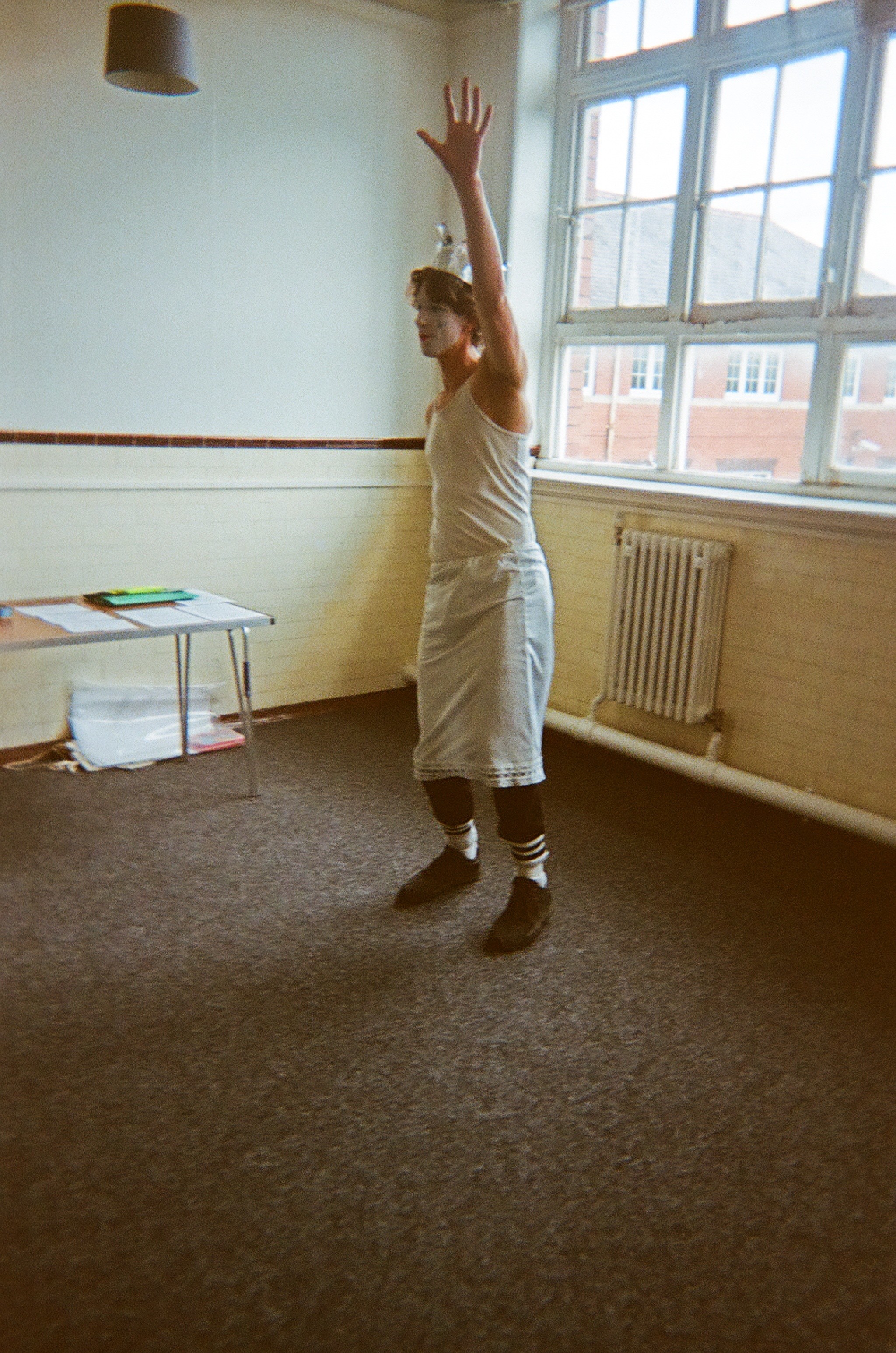 A person indoors, standing on a carpeted floor near a large window with multiple panes, raising one arm upward. The individual wears a white tank top, a light-colored wrap or skirt over black pants, striped socks, and dark shoes. A radiator and a table with papers and a green object are visible in the room, which has cream-colored walls with a dark trim and a hanging light fixture.