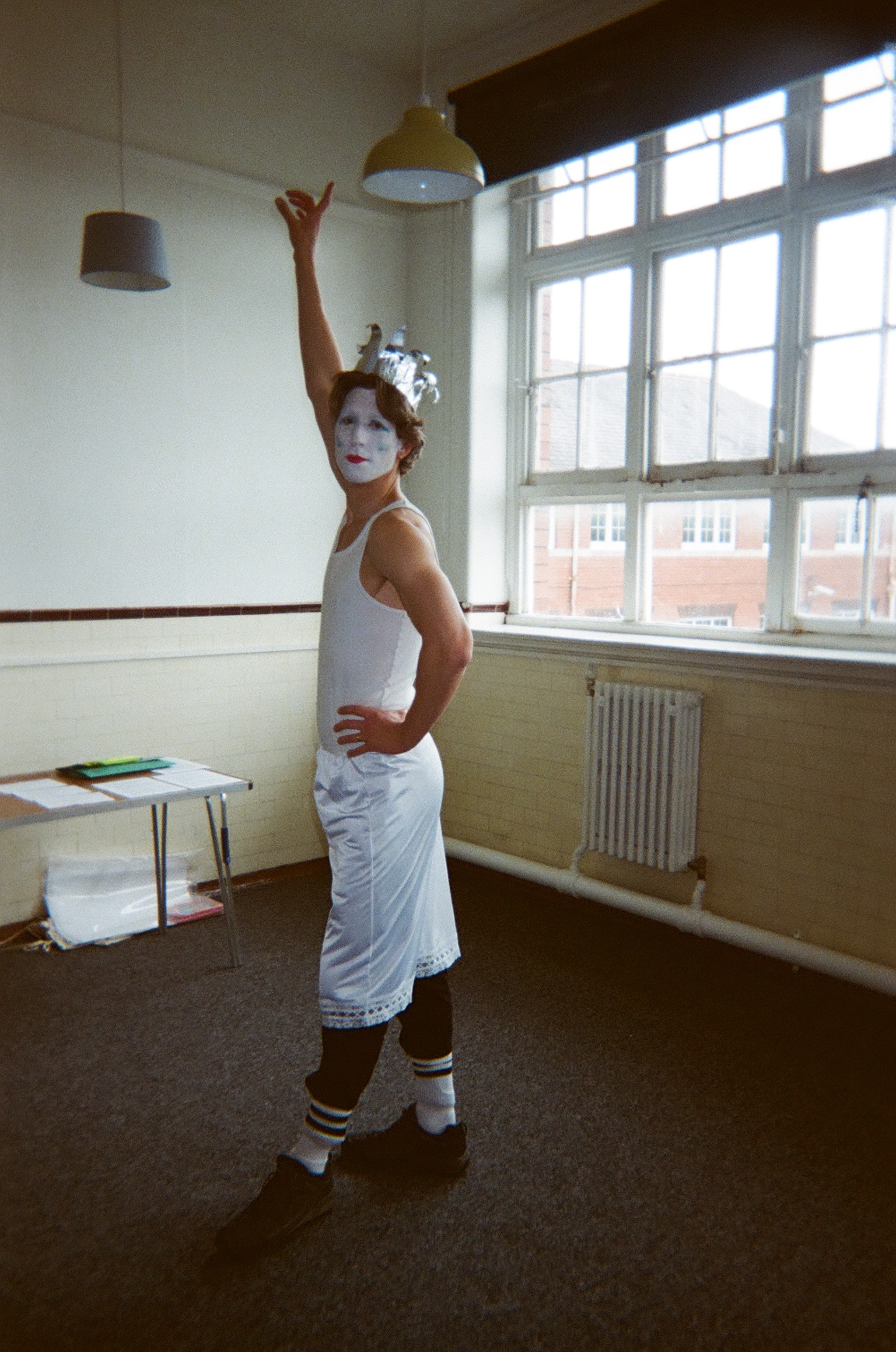Person with white face paint and a silver paper crown, wearing a white sleeveless top, white skirt over black leggings, and striped socks, posing with one arm raised and the other on their hip in a room with large windows and two ceiling lights.