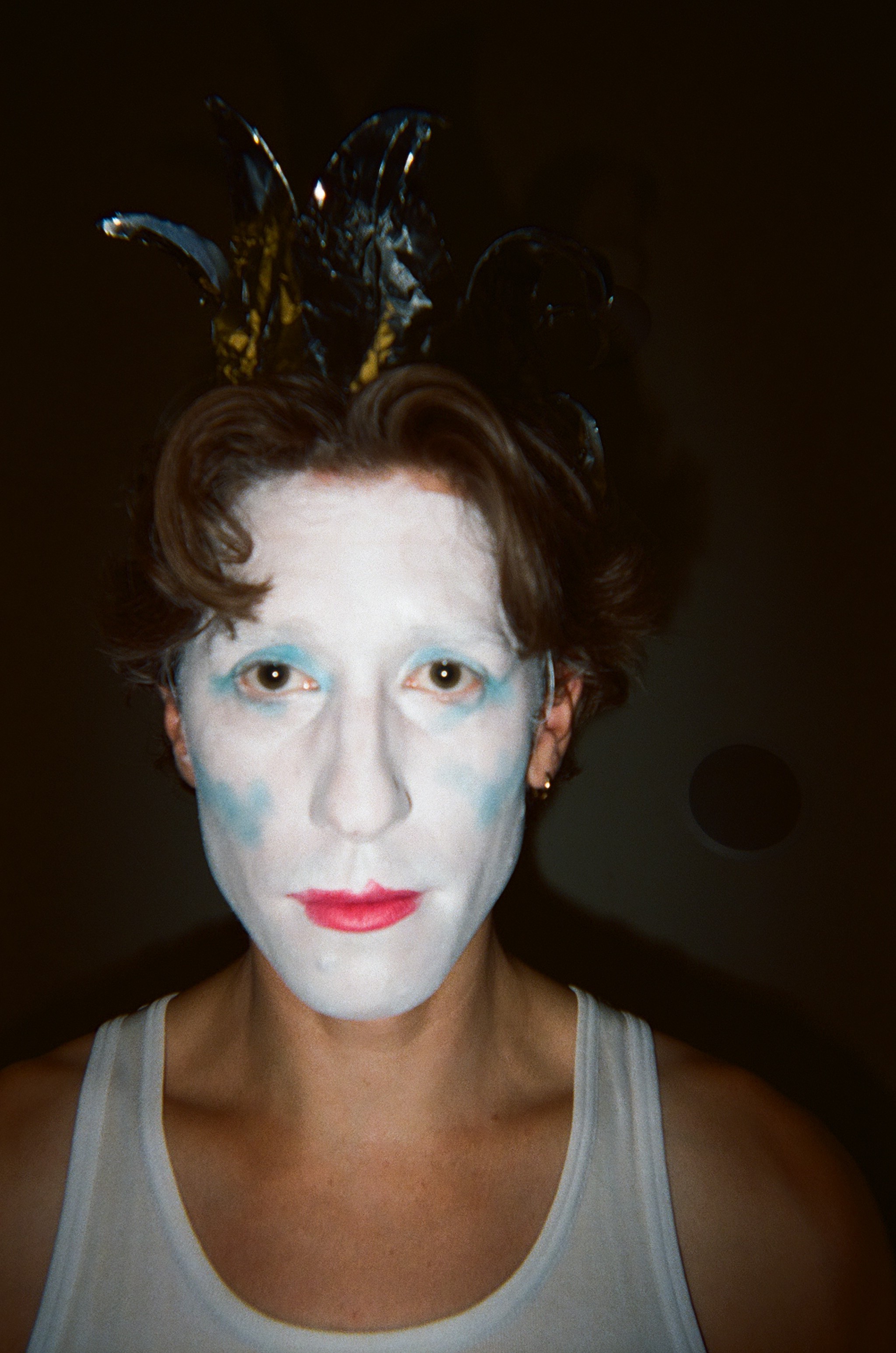 Person with white face makeup, blue eyeshadow, red lips, and short brown hair wearing a black and gold jester-style headpiece and a white tank top against a dark background.