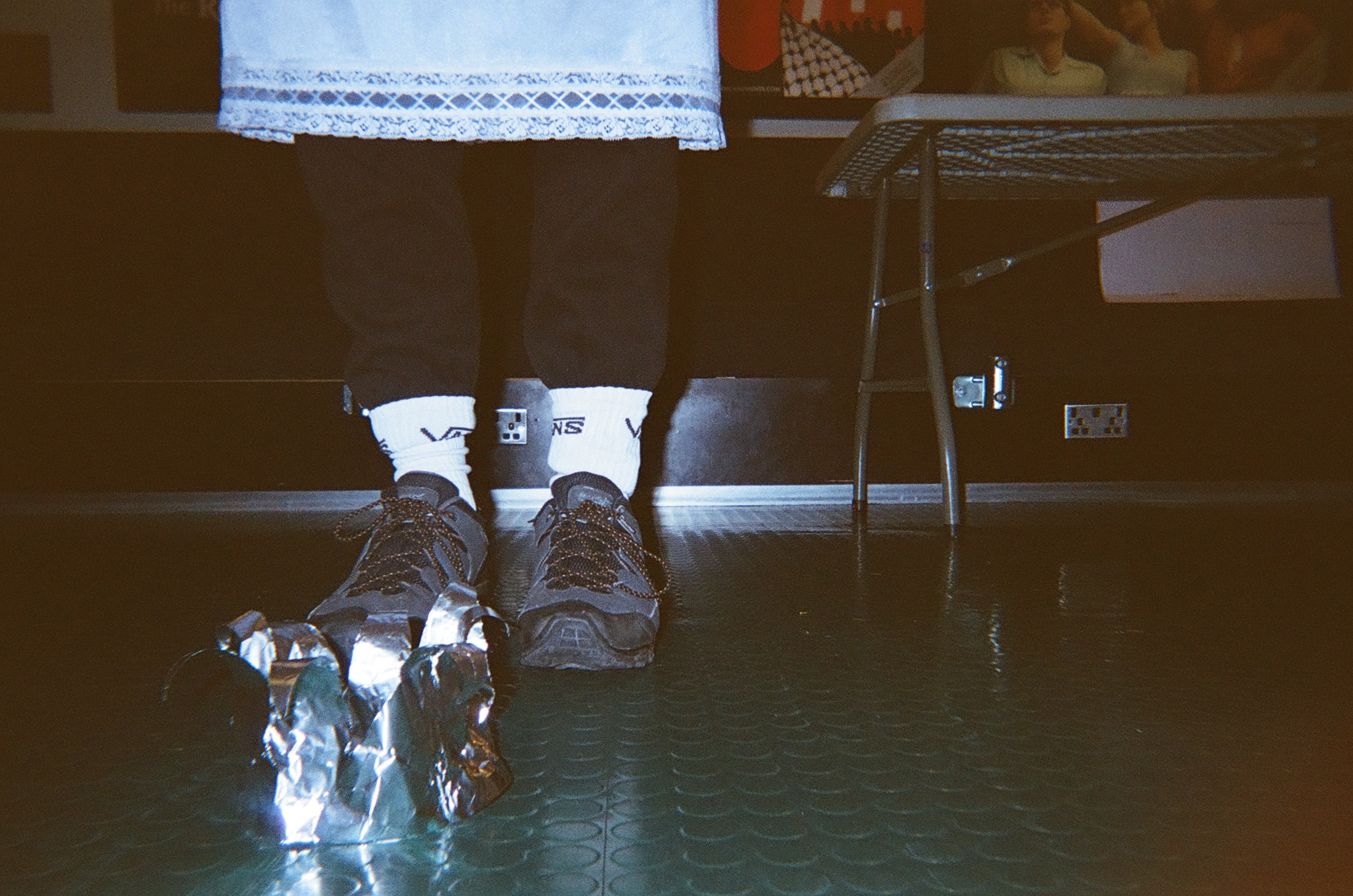 Close-up of a person standing on a textured floor wearing black shoes with bulky silver wrapped material attached to the left shoe, white Vans socks, and black pants, with part of a table visible on the right side.