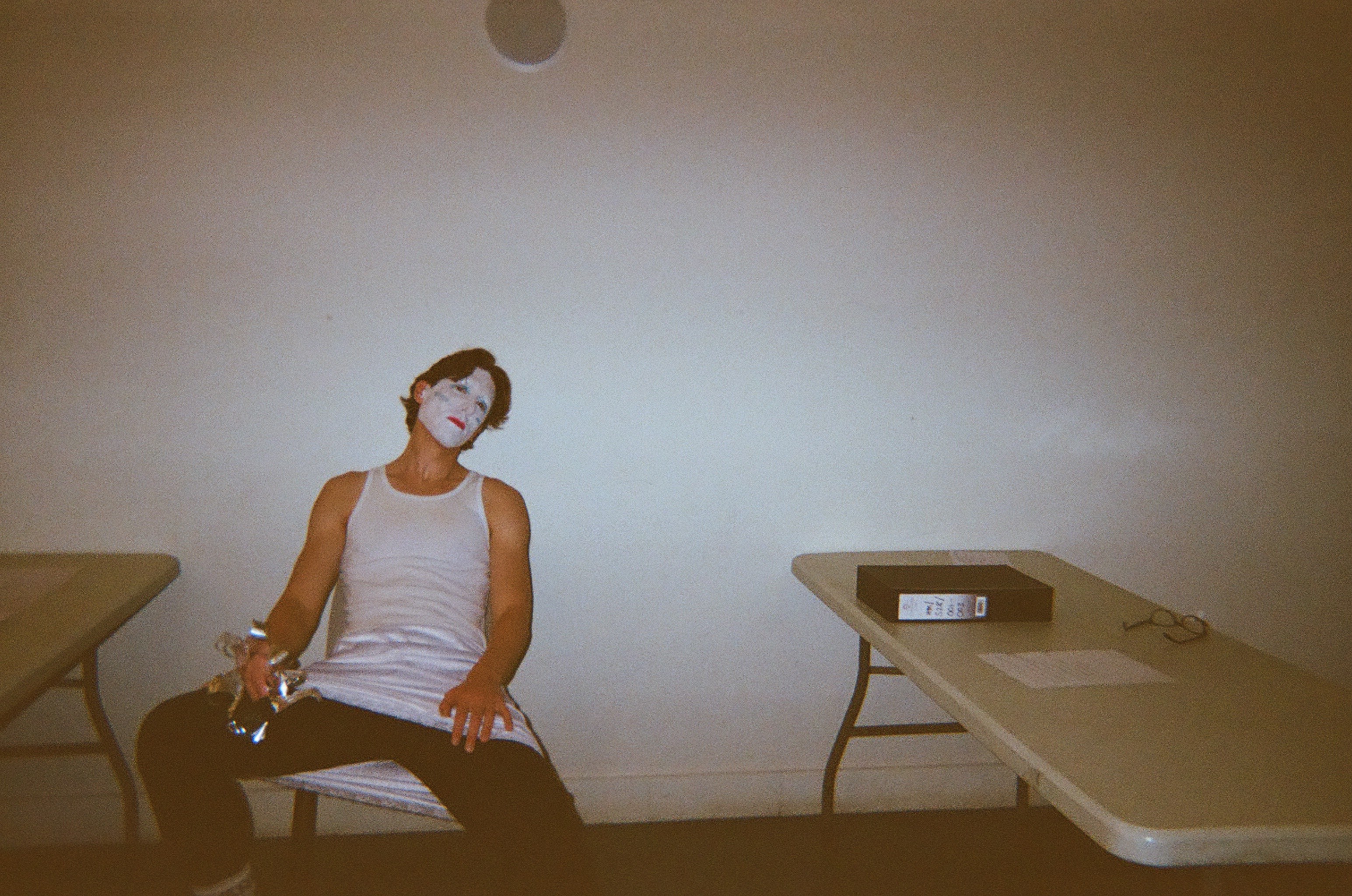 Man with white face paint sitting slouched on a chair between two folding tables in a dimly lit room, holding a crumpled foil object in one hand, with glasses and a binder on the right table.