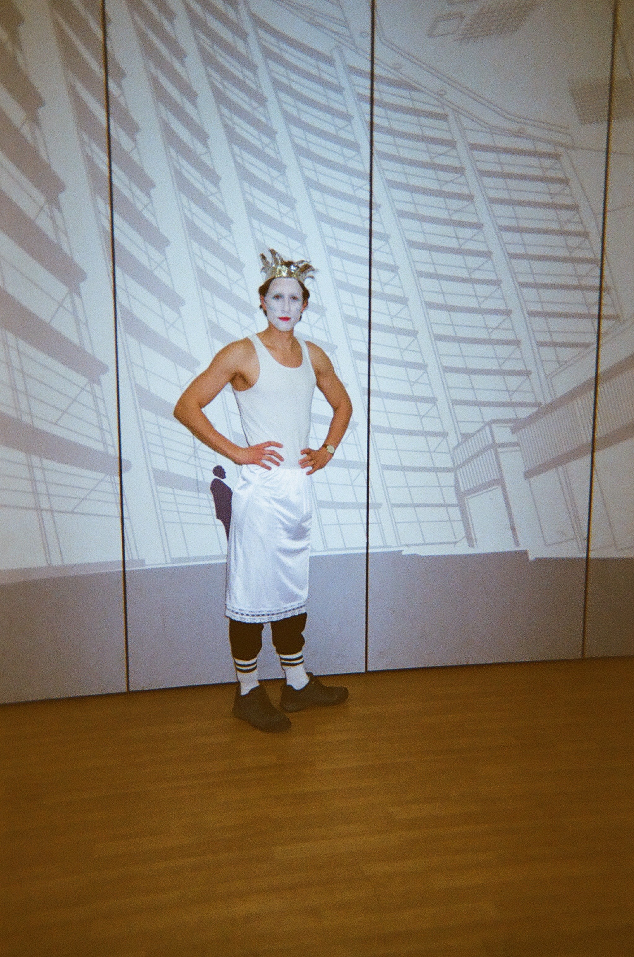 Person with white face paint wearing a crown, white tank top, white apron over black pants, striped socks, and dark shoes standing with hands on hips against a backdrop of architectural line drawings.
