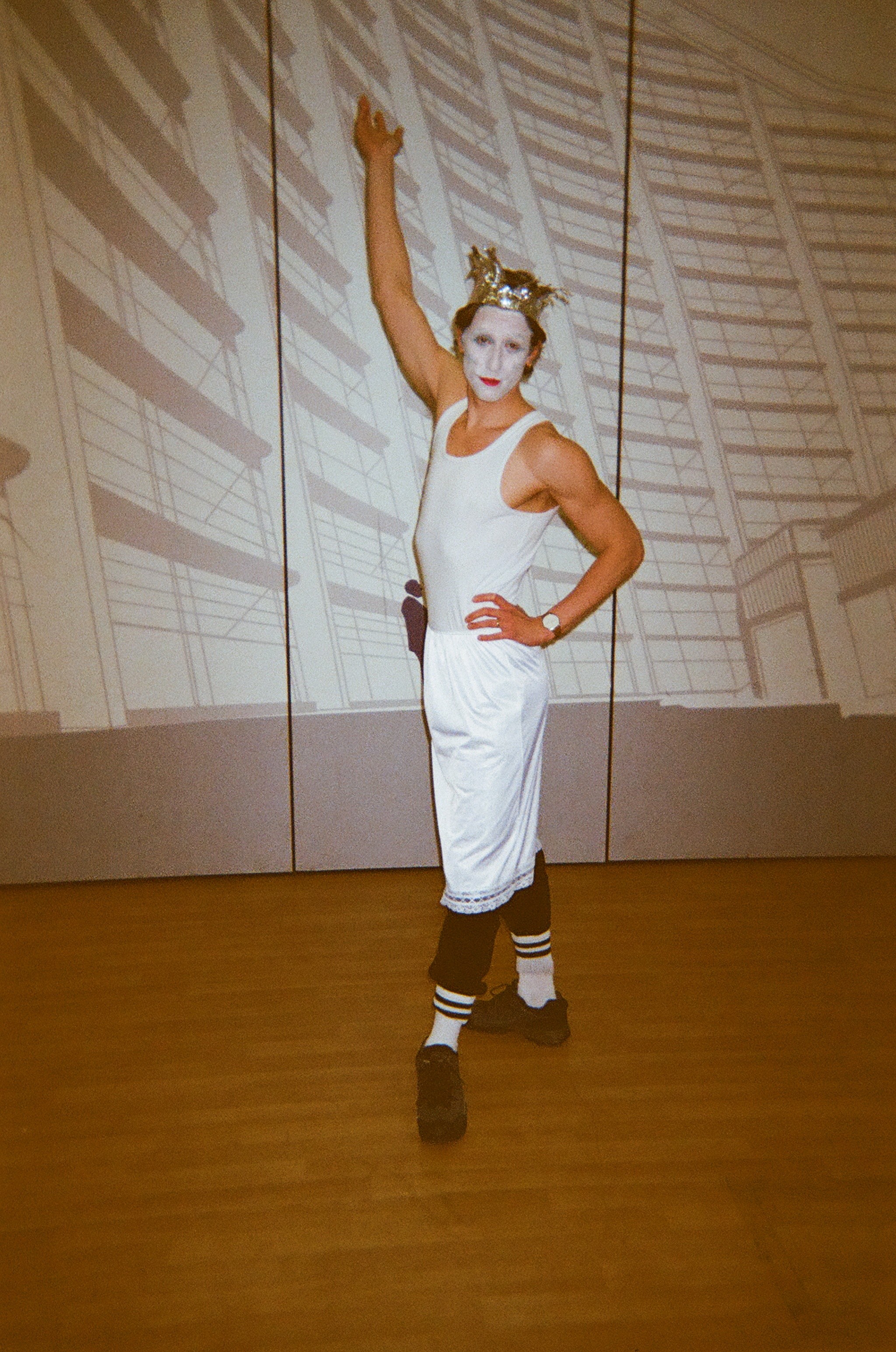 Person dressed in a white sleeveless dress over black leggings and black shoes, wearing striped socks and a gold crown, with face painted white and red lipstick, posing with one arm raised and the other hand on their hip against a background with abstract shadow patterns.