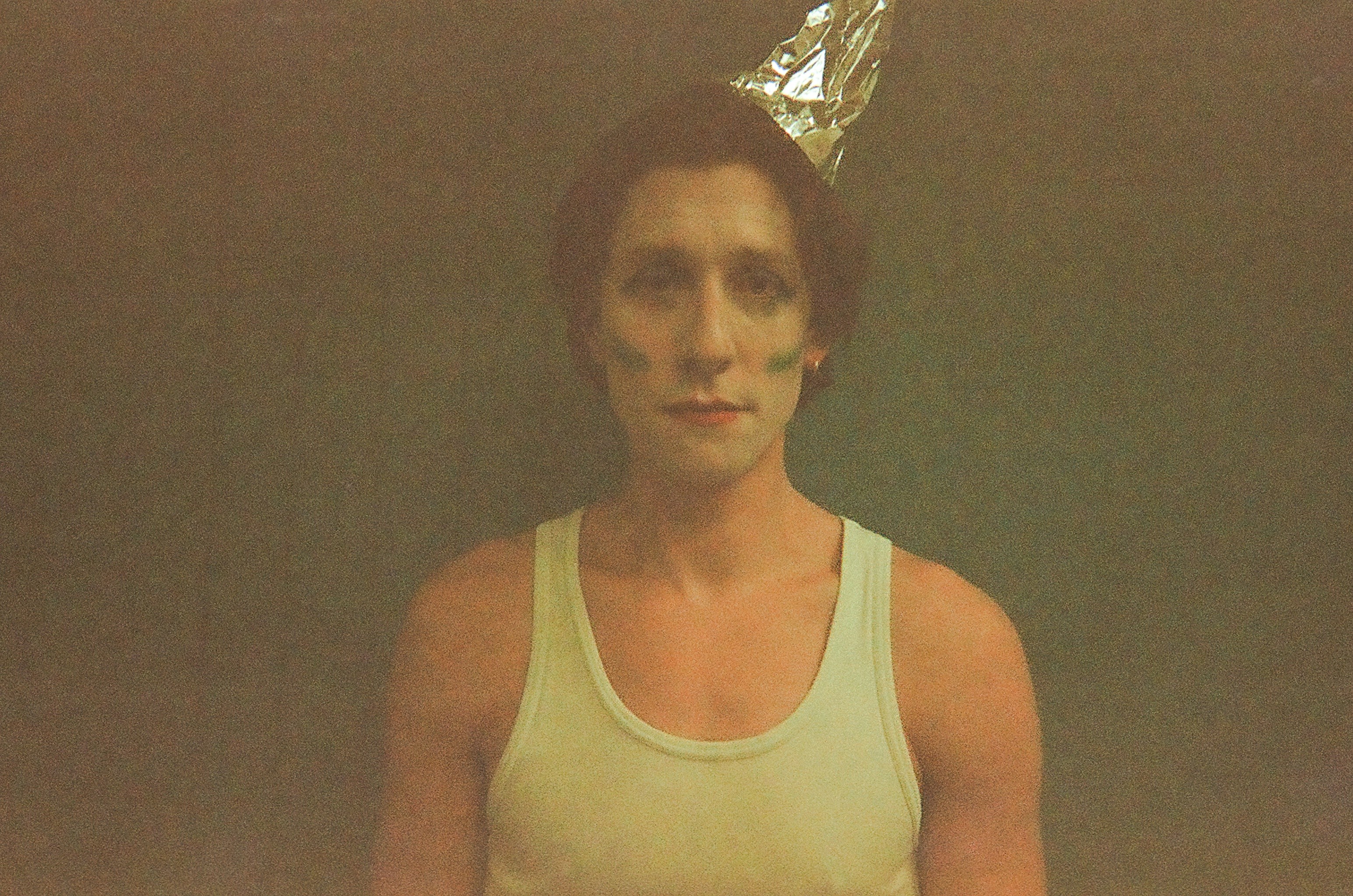Person wearing a white tank top with green face paint on their cheeks and a cone-shaped hat made of crumpled aluminum foil, standing against a muted green background.
