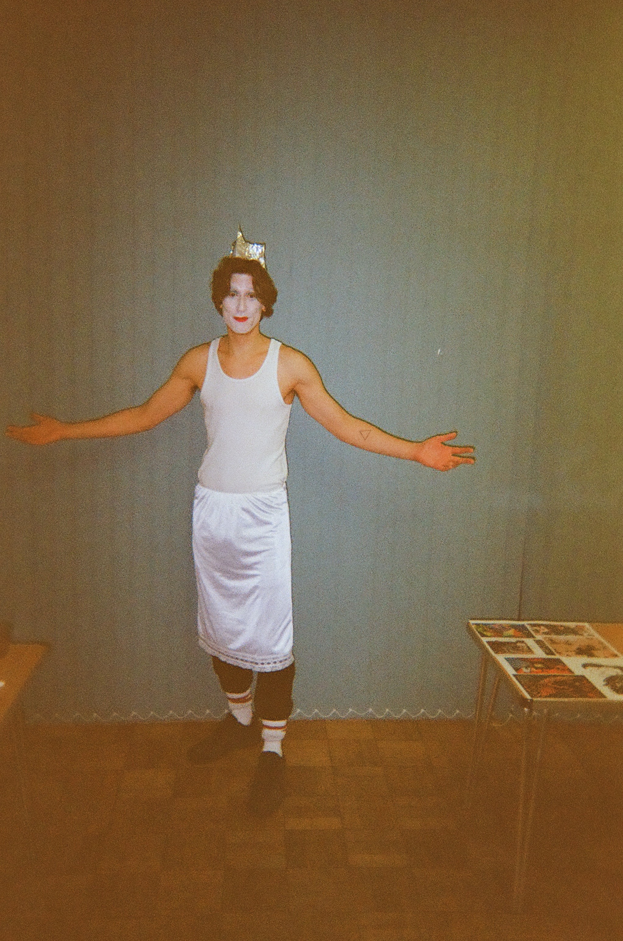 Person wearing a white tank top, white satin skirt over black pants, white socks with stripes, black shoes, and a silver crown, standing with arms open in a room with a wooden floor and grey curtains, next to tables with magazines.