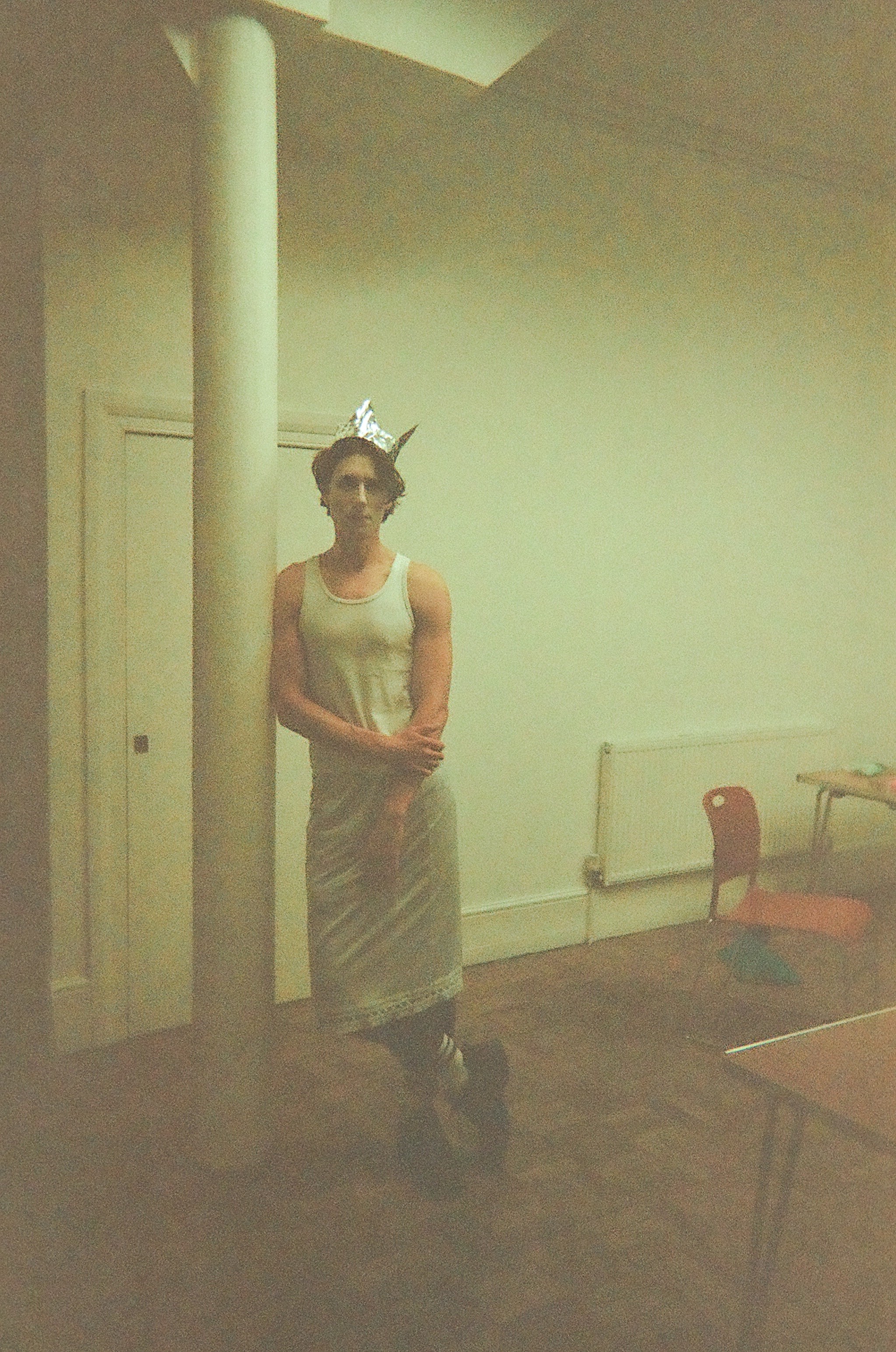 Young person standing indoors leaning against a white pillar, wearing a sleeveless dress, a makeshift crown made from aluminum foil, and striped socks with boots, in a dimly lit room with a red chair and radiator in the background.