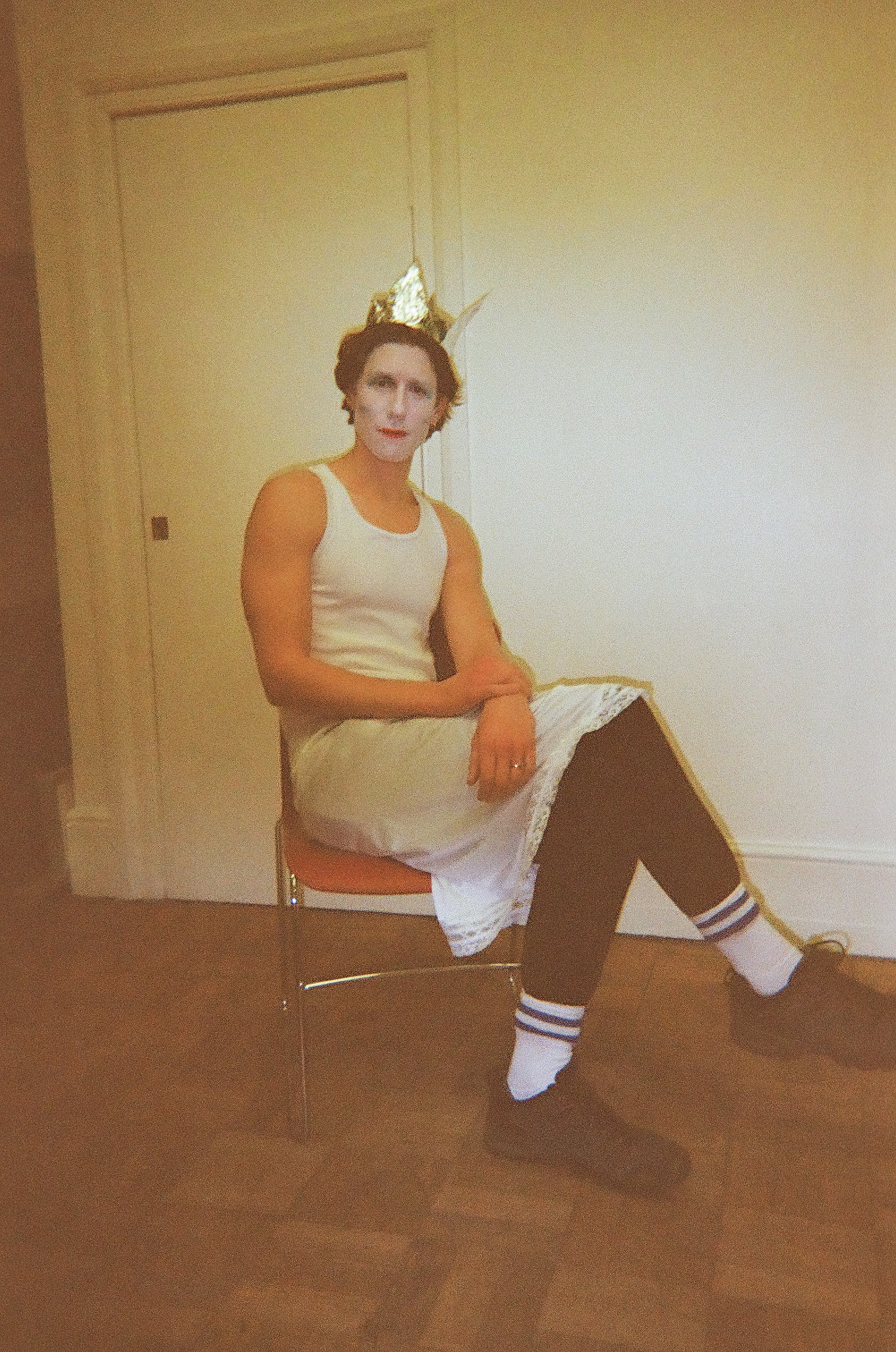 A person sitting on a chair indoors wearing a white sleeveless dress over black leggings, white socks with blue stripes, and black shoes, with a gold crown on their head and white face makeup.