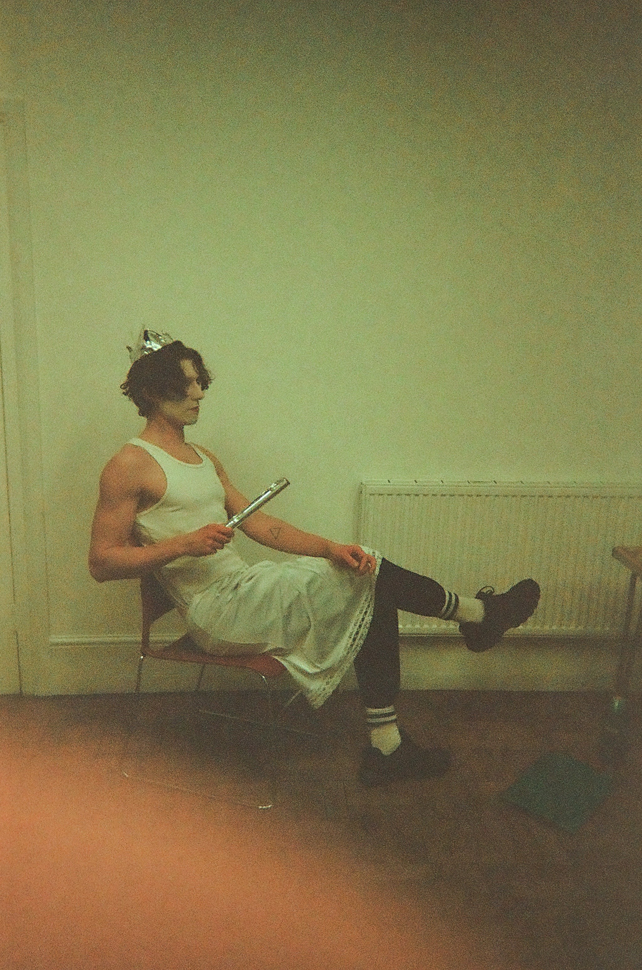 Person with curly hair and white face paint wearing a silver paper crown, white tank top, white skirt, black leggings, striped white socks, and black shoes sitting sideways on a chair with one leg crossed over the other, holding a silver flute.