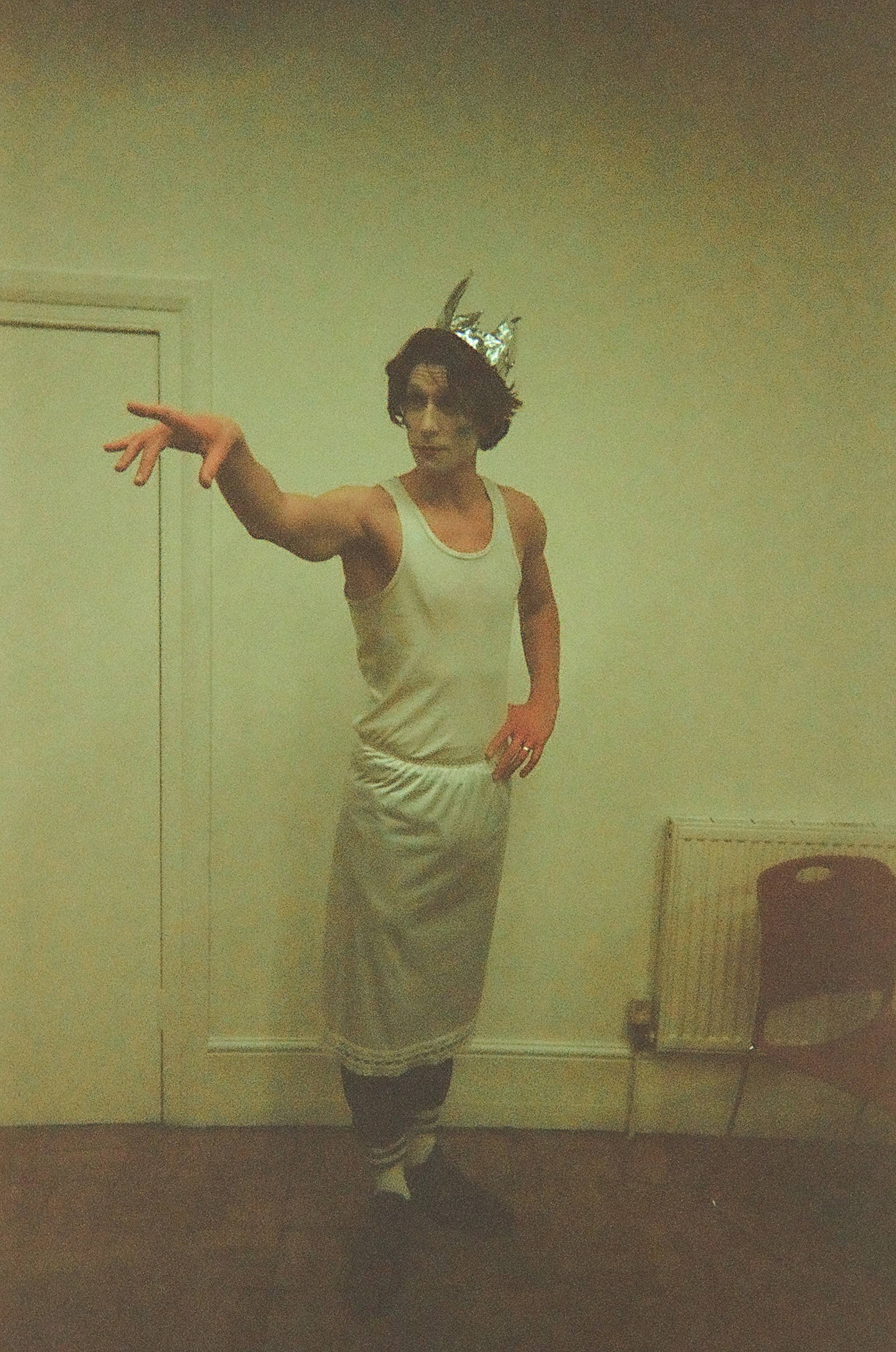 Person wearing a silver foil crown, white tank top, and long white skirt posing dramatically with one arm extended and the other on their hip in a simple room with a door, radiator, and empty chair.