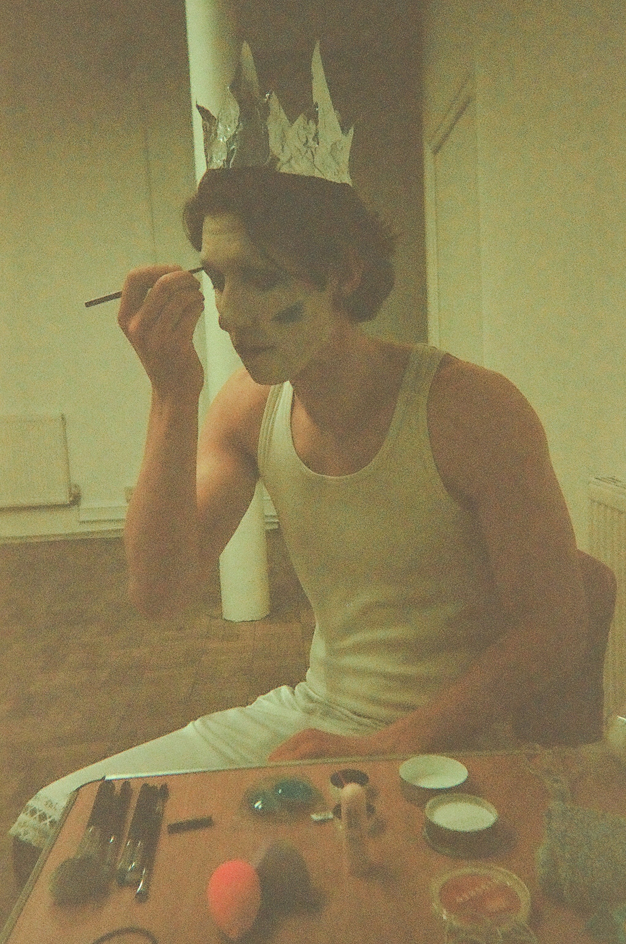 Person wearing a white tank top and a paper crown applies makeup using a brush while seated at a table with various makeup items.