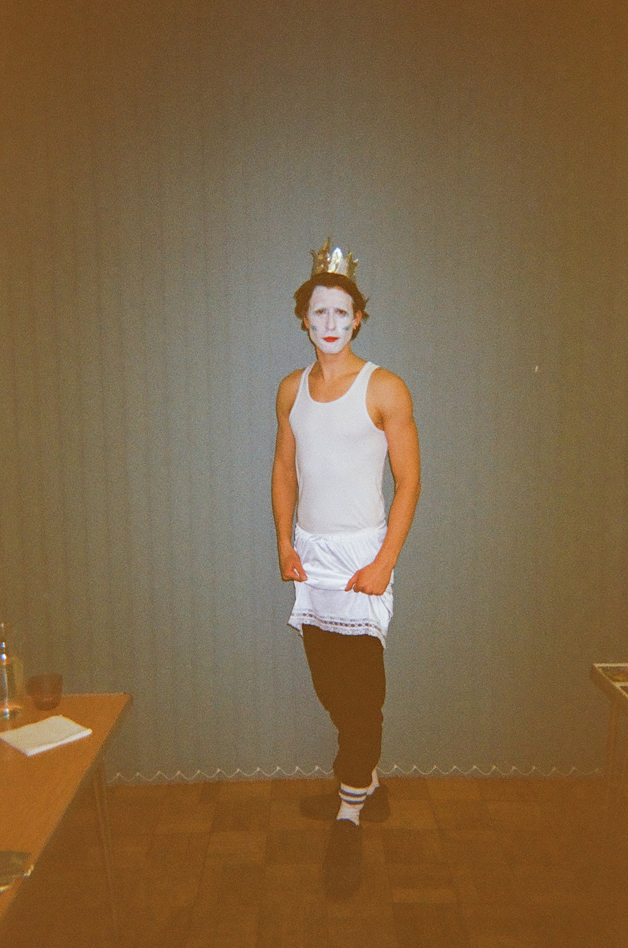 Person standing indoors against a plain wall, wearing a white tank top, a white skirt-like garment over black pants, black shoes, and white socks with black stripes, with a gold crown on their head and face painted white with red lips and blueish markings on the cheeks.