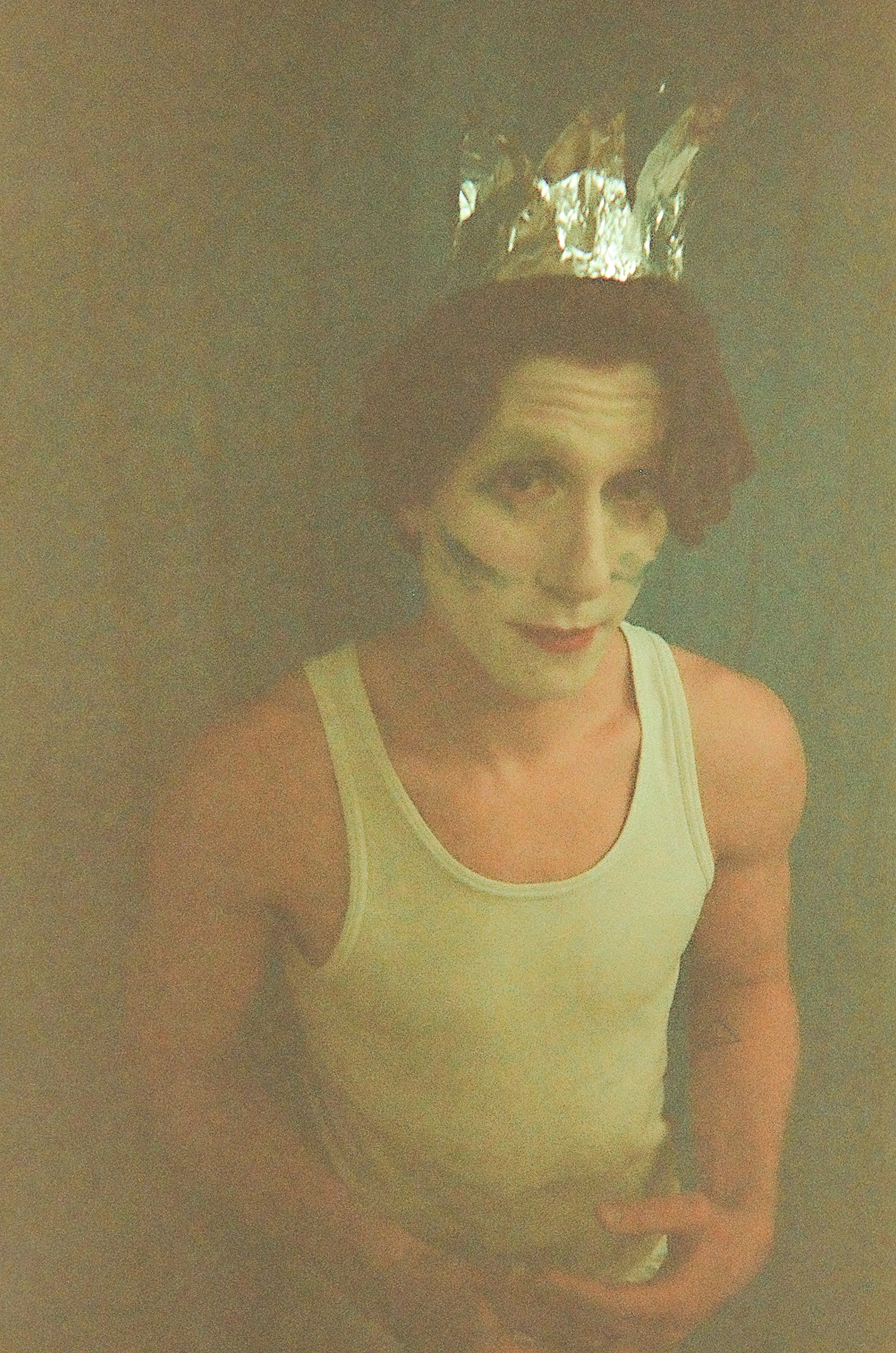 Person with clown-like face paint wearing a silver crown and a white sleeveless shirt, posing against a blue background.