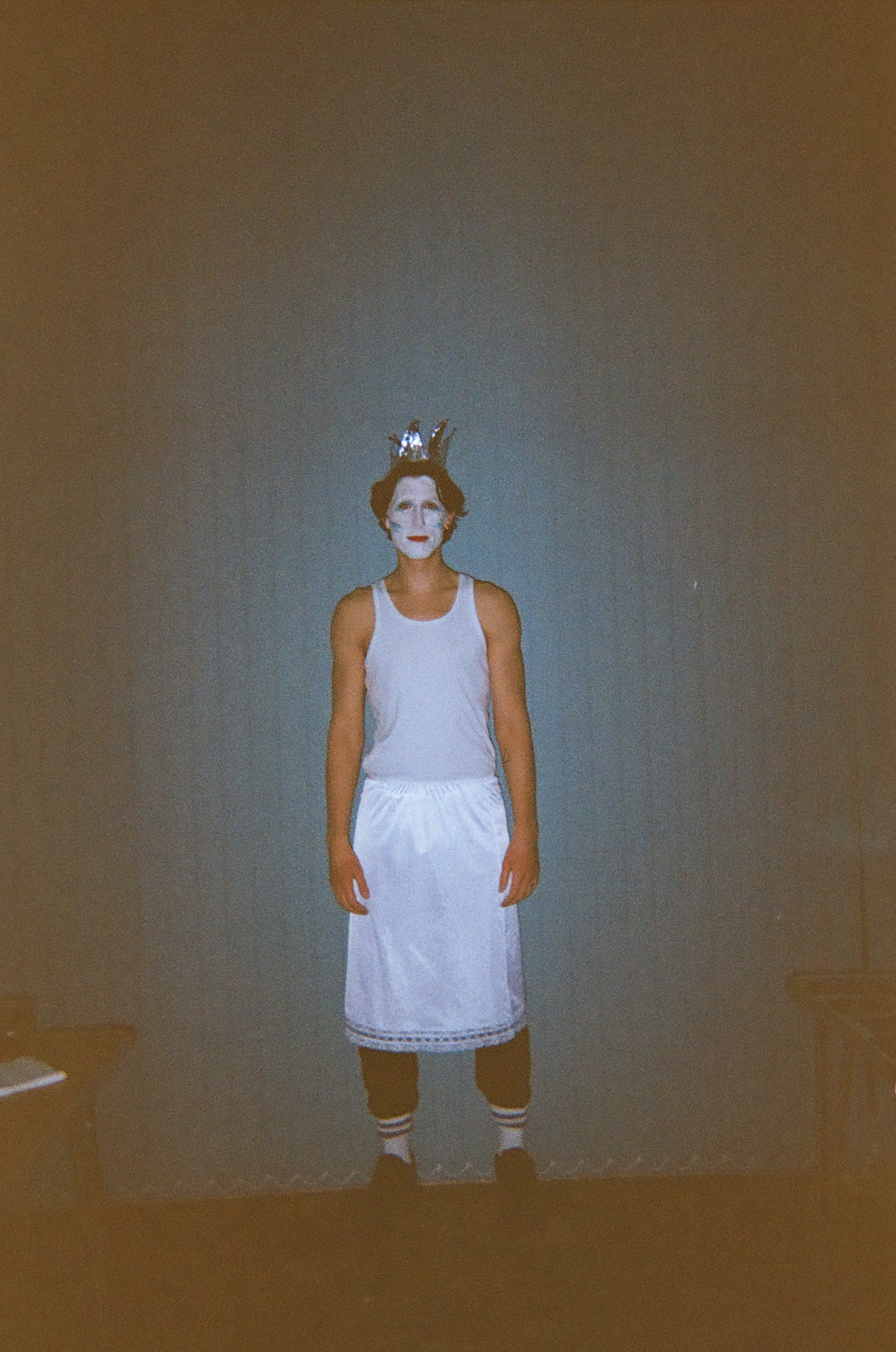 Person standing indoors against a plain wall wearing a white tank top, a white skirt, black pants underneath, striped socks, and black shoes, with face painted white and a silver crown on their head.