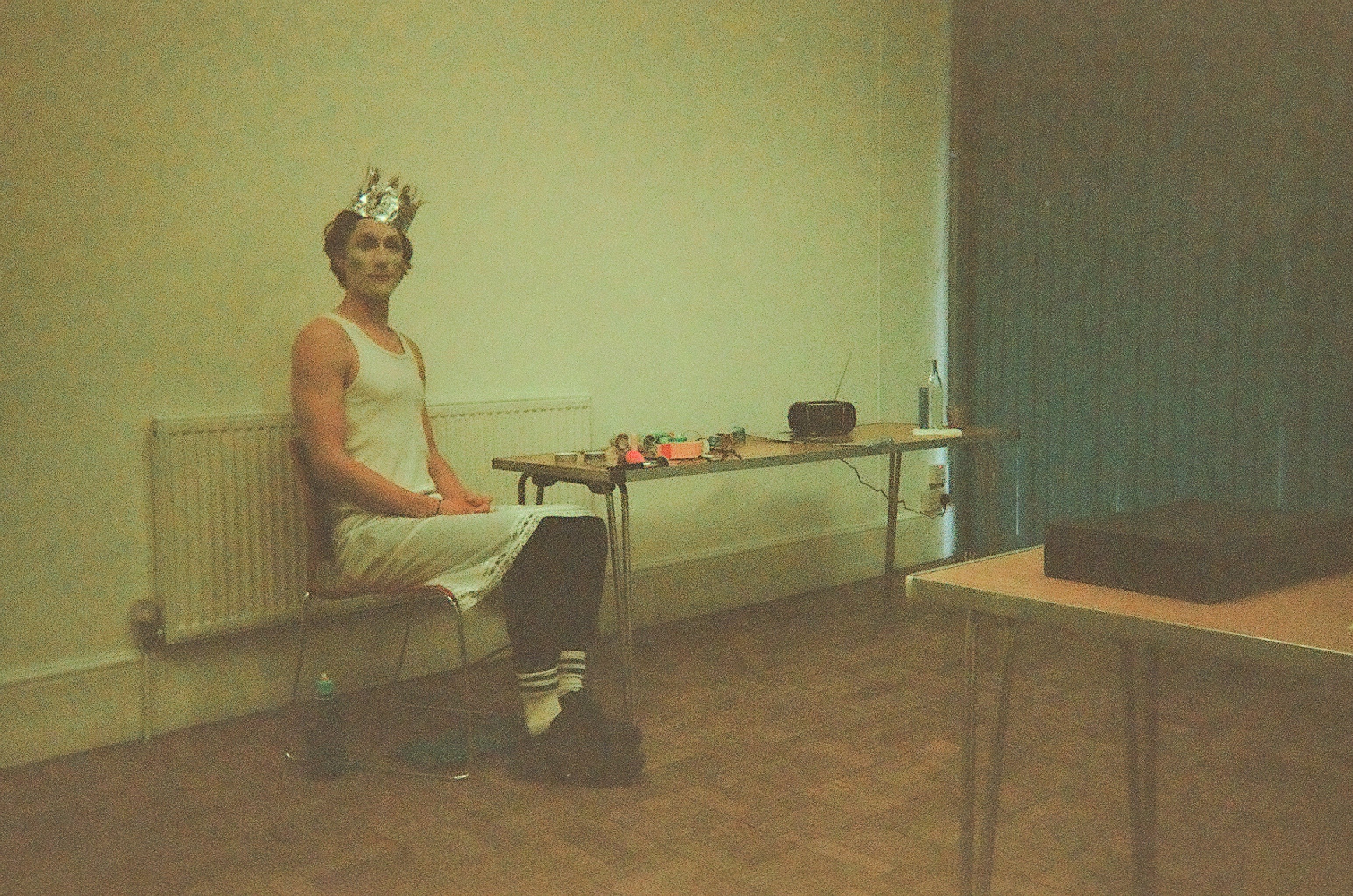 Person wearing white dress, striped socks, dark shoes, and a shiny crown, sitting on a chair next to a table with random items, in a sparsely furnished room with beige walls and a heater behind them.
