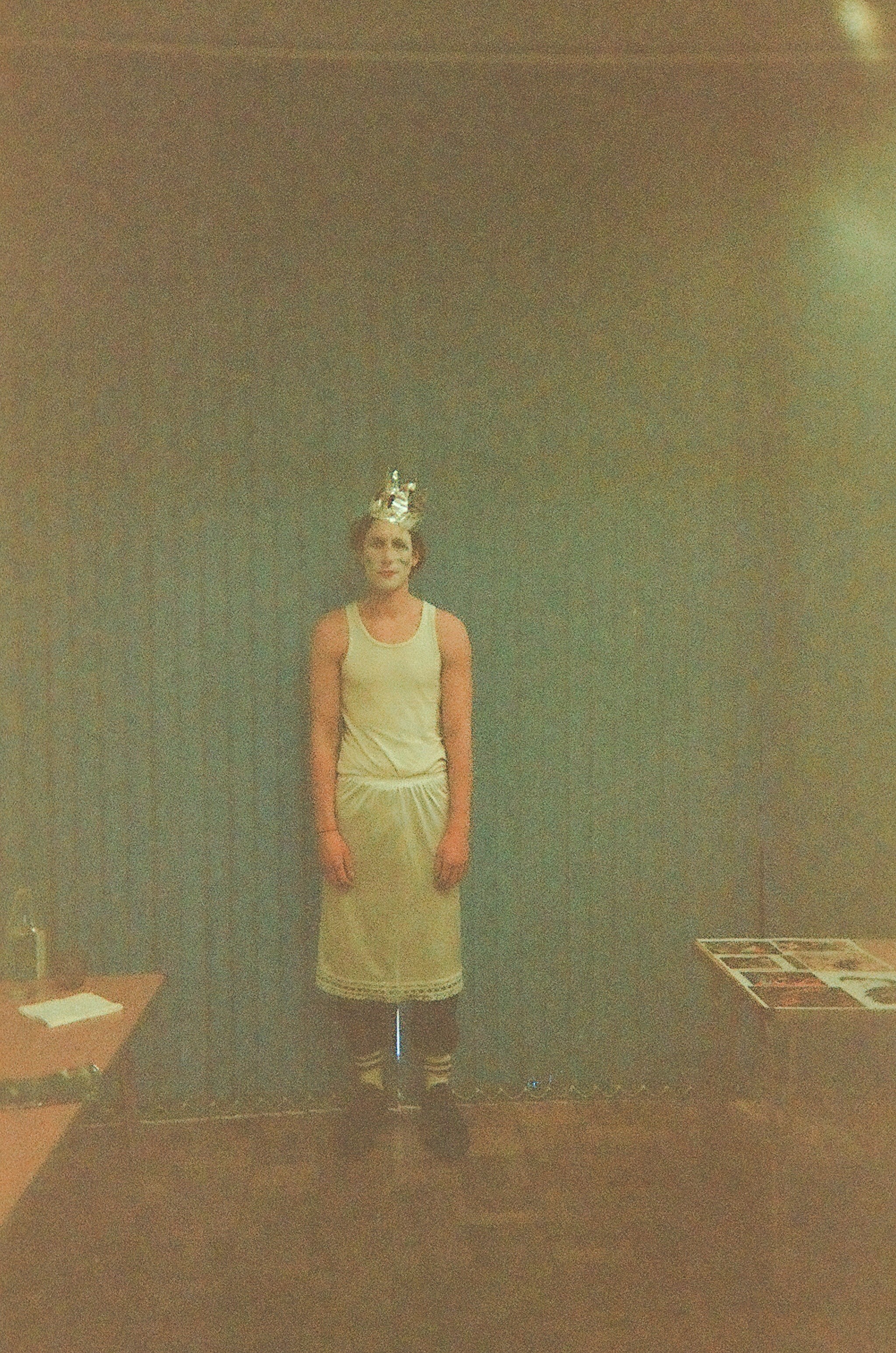 Person standing against a dark curtain wearing a white sleeveless shirt, a white skirt, striped socks, black shoes, and a silver crown headpiece.