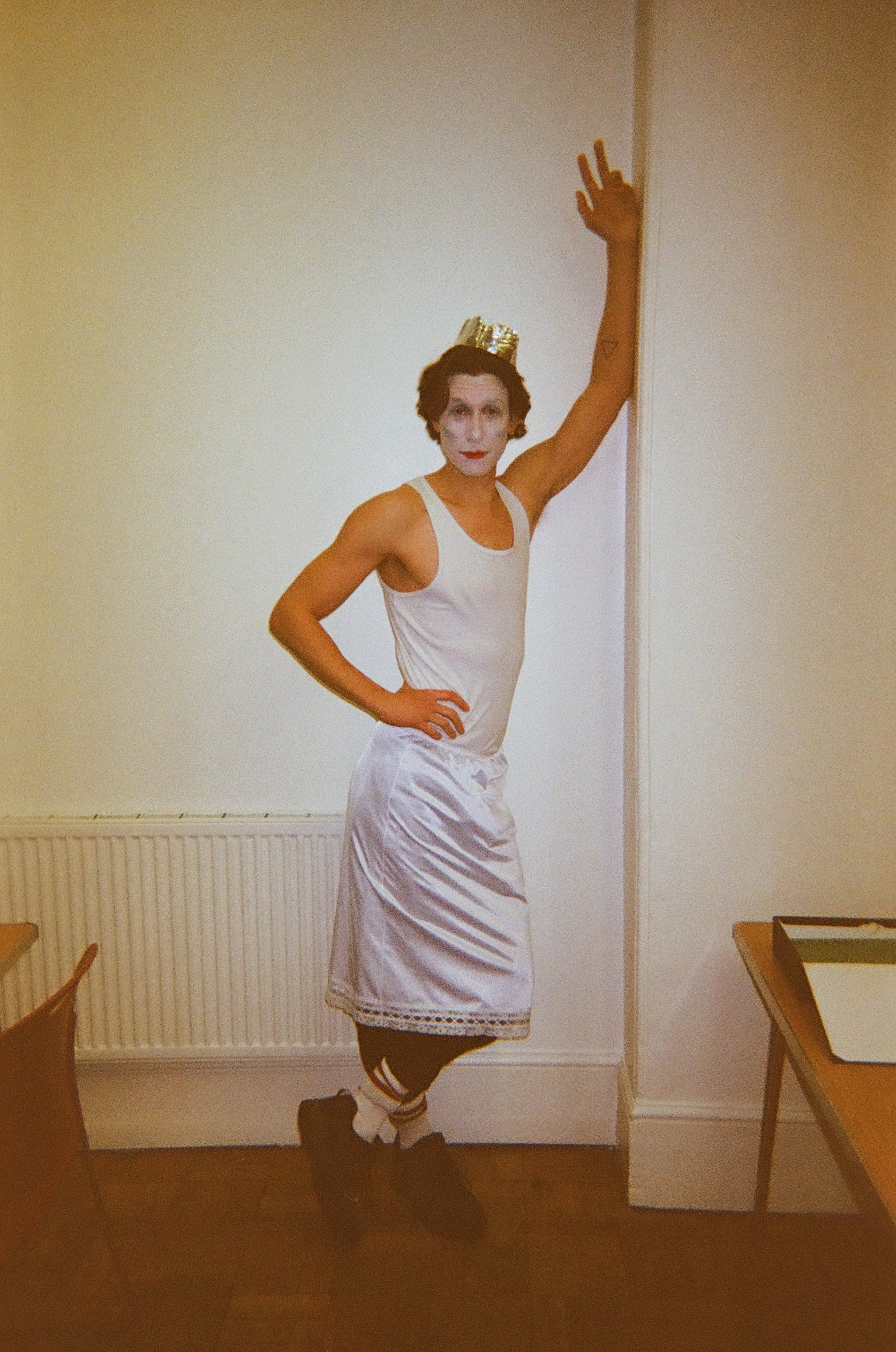 Person wearing a gold crown, white face paint with red lips, a white tank top, a shiny white skirt, black leggings with stripes, white socks, and black shoes, standing indoors against a cream wall with one arm raised and resting on the wall.