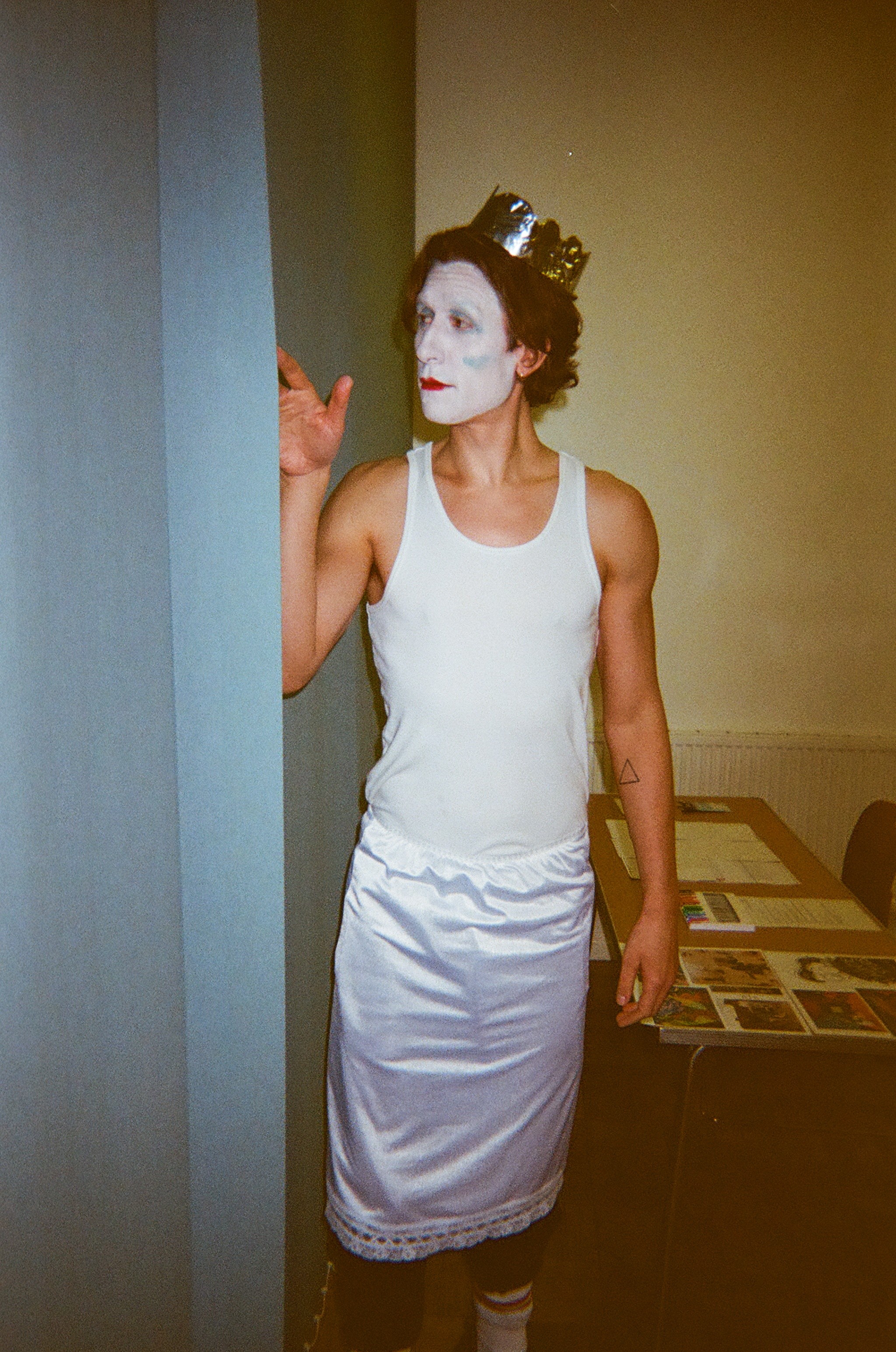 Person wearing white face paint with red lips and a metallic crown, dressed in a white tank top and shiny white skirt, standing beside a wall with one hand raised.