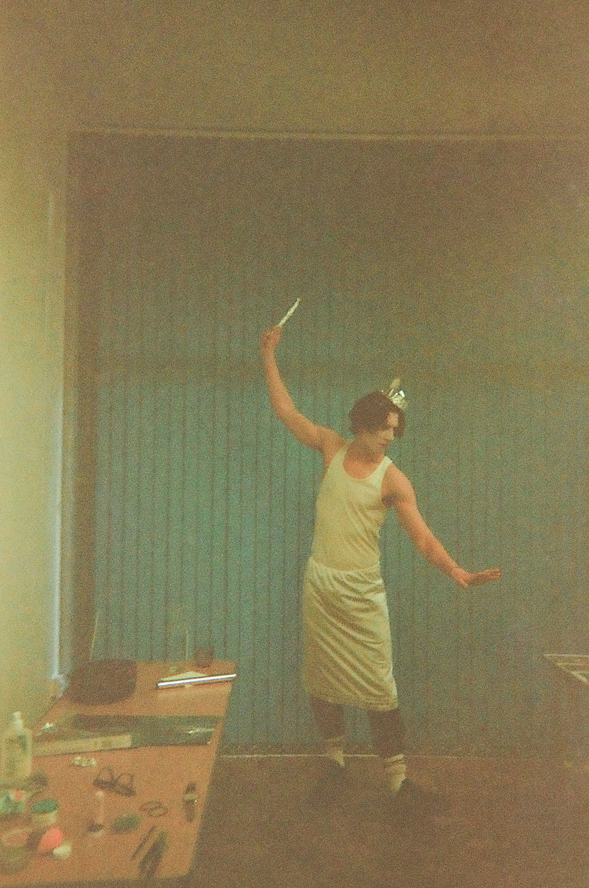 Person wearing a crown and a beige tank top with a skirt-like garment, striking a pose with one arm extended and the other holding an object, standing in a dimly lit room with a blue curtain backdrop and a cluttered table with various items nearby.