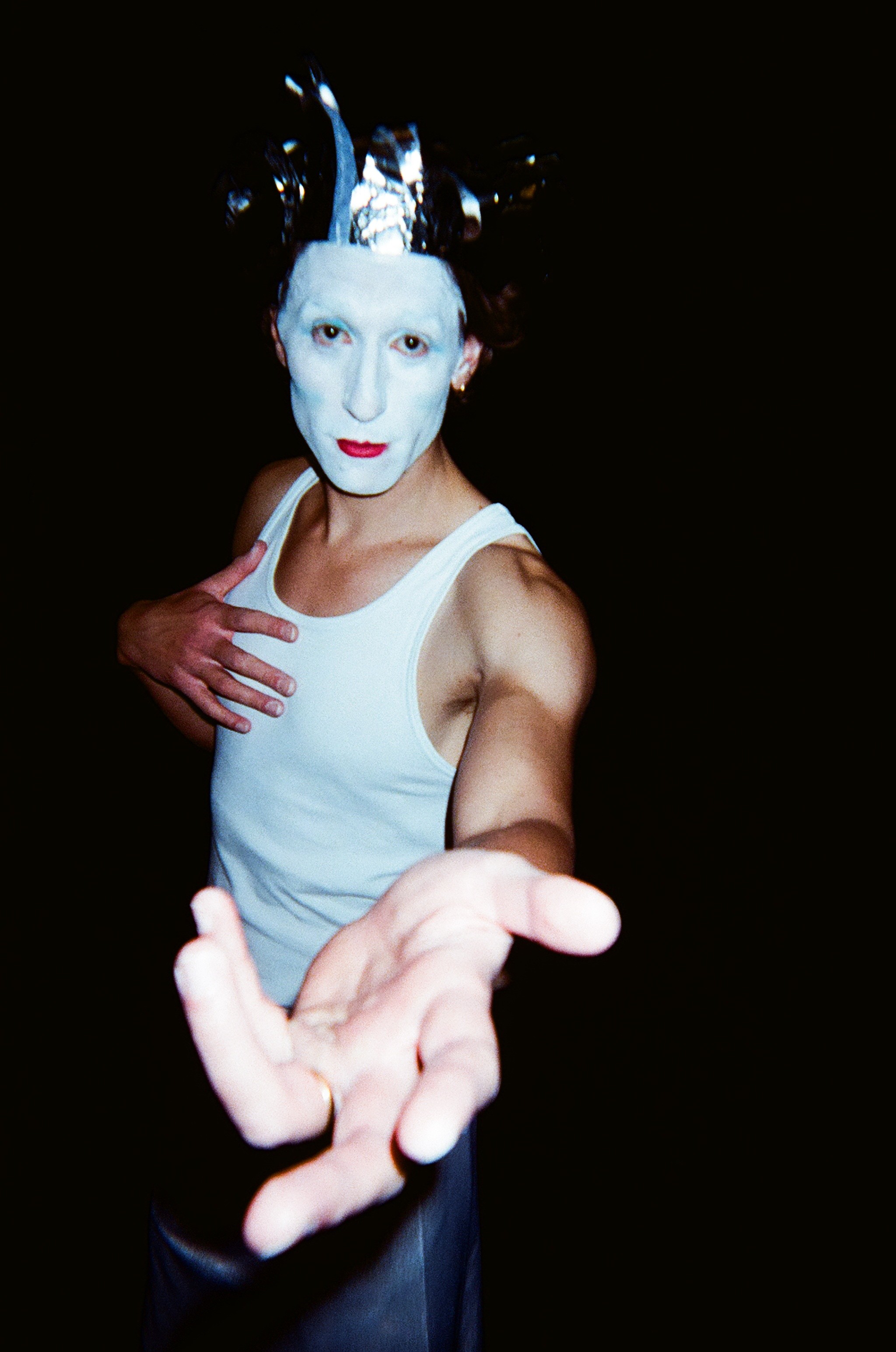 Person with white face paint, red lipstick, and a black and white headpiece, wearing a white tank top, reaching out with one hand toward the camera against a black background.