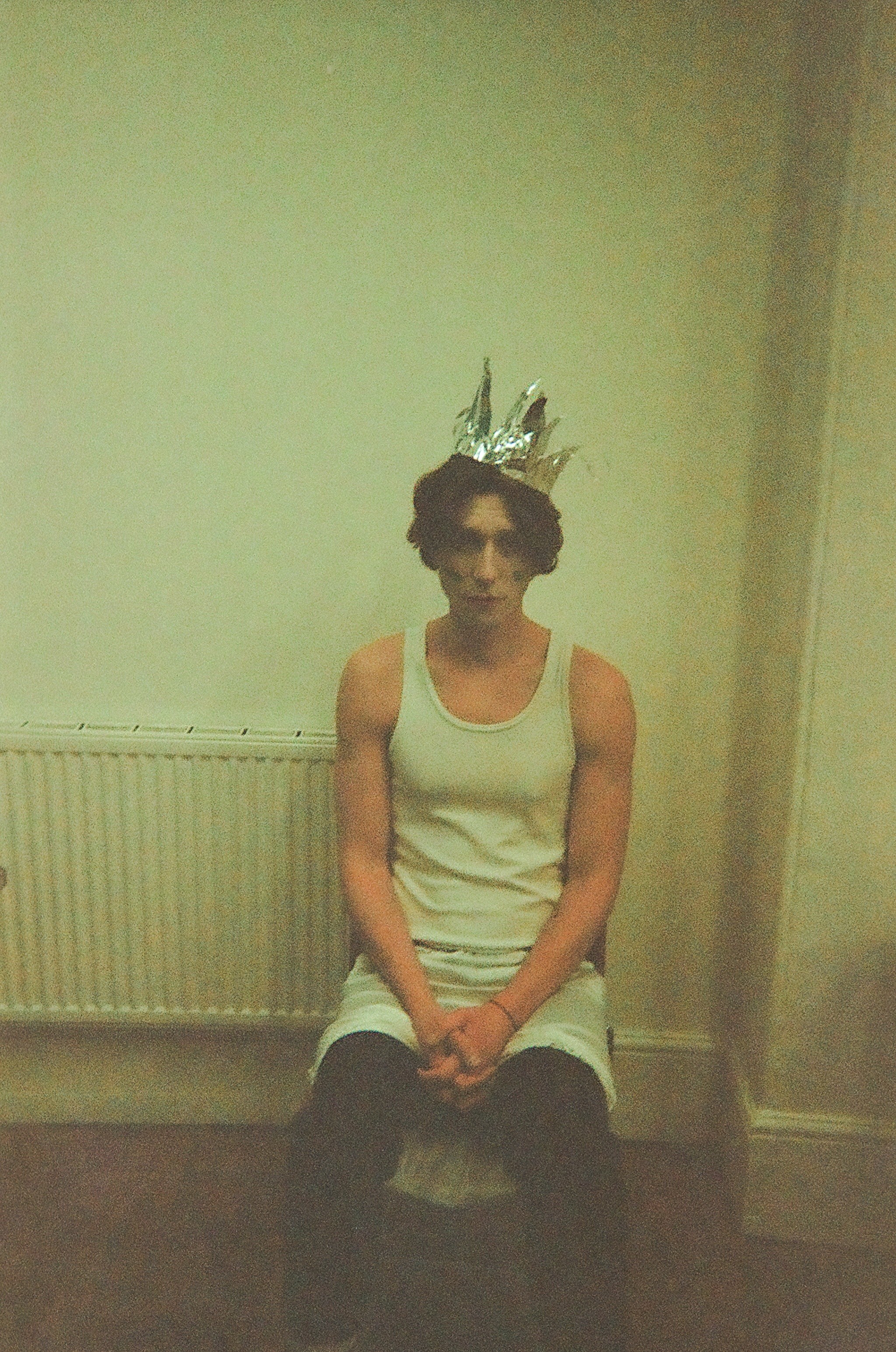 A young person with curly hair sits on a chair against a plain wall, wearing a metallic crown, a white sleeveless top, and dark pants, with hands clasped.