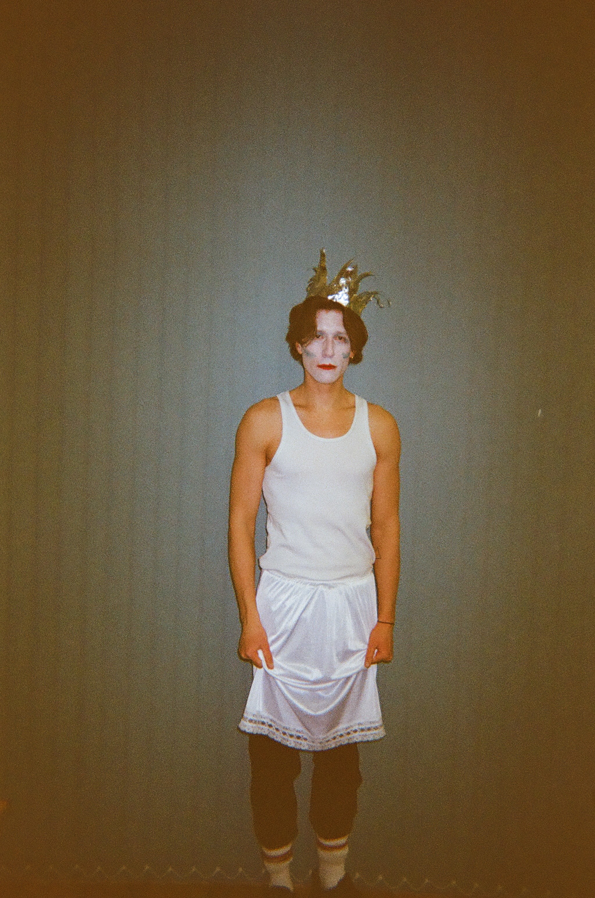 Person wearing a white tank top, white skirt with decorative trim, black pants, striped socks, and a shiny gold crown with points, standing against a plain wall with a neutral expression and white face makeup with red lips.