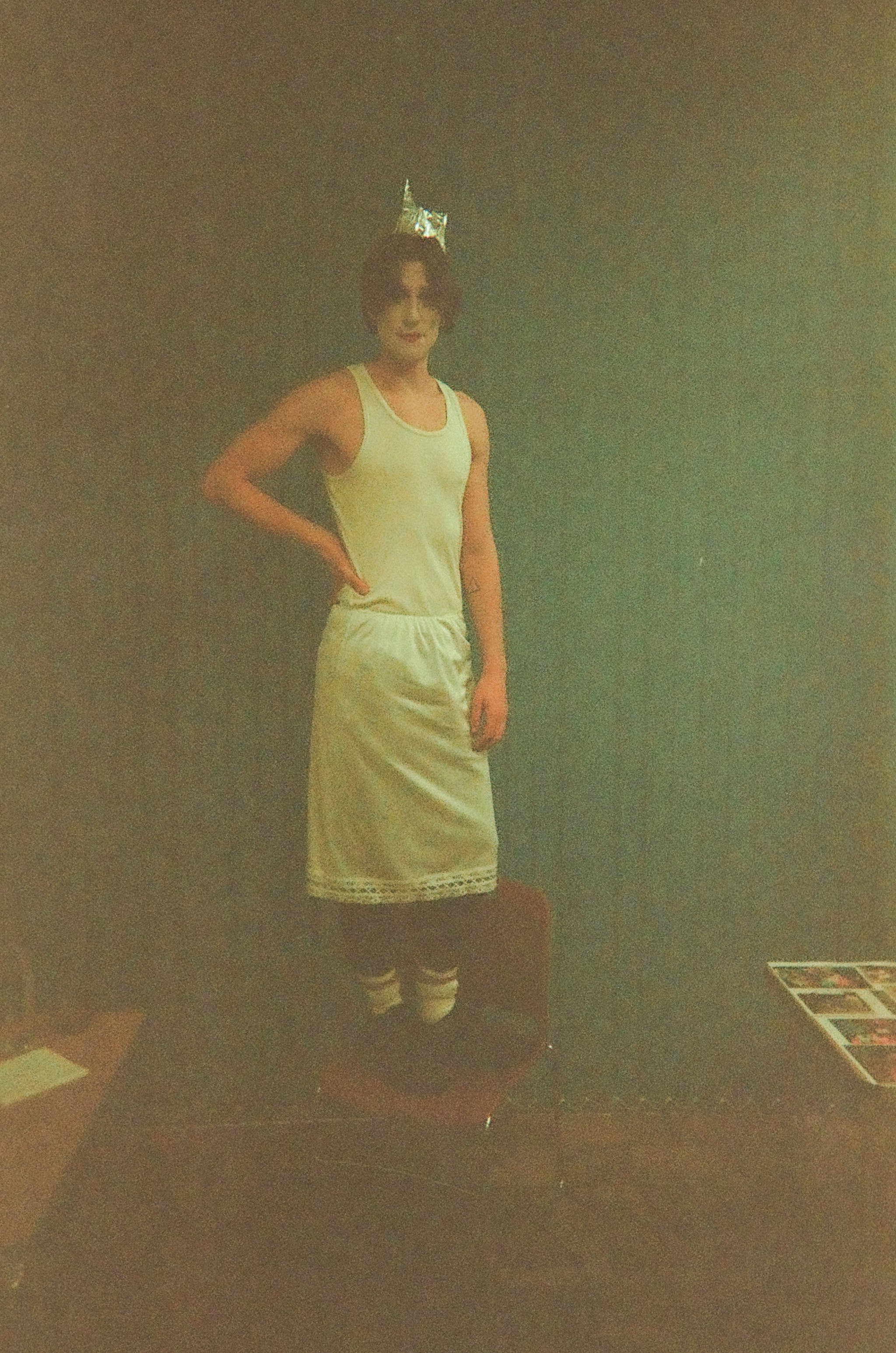Person wearing white tank top, a white skirt with lace trim, white socks, black shoes, and a small foil crown standing on a chair against a teal wall.