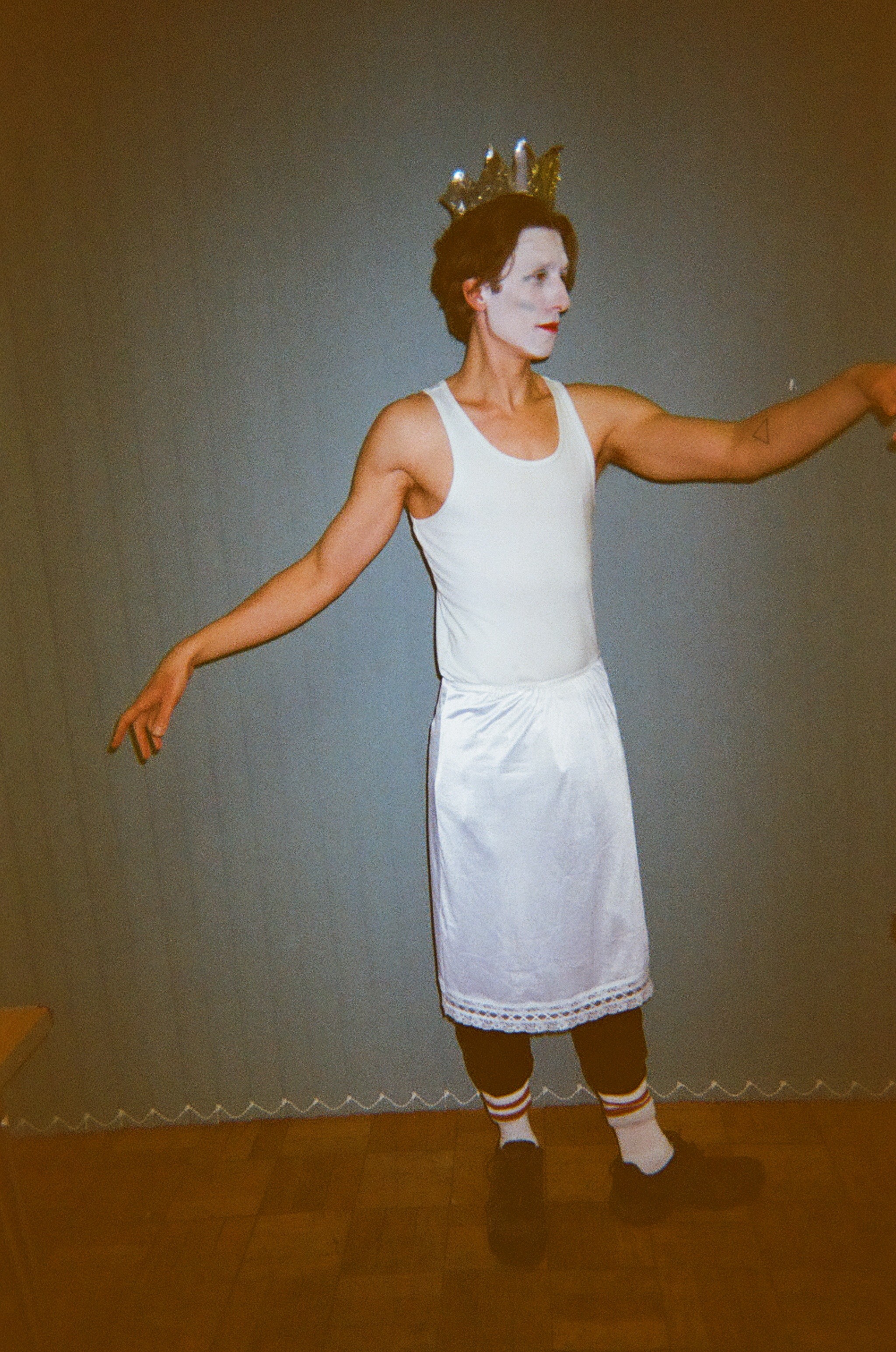 Person wearing white face paint, a gold crown, white tank top, white skirt with lace trim, and striped socks, standing with arms extended against a plain wall.