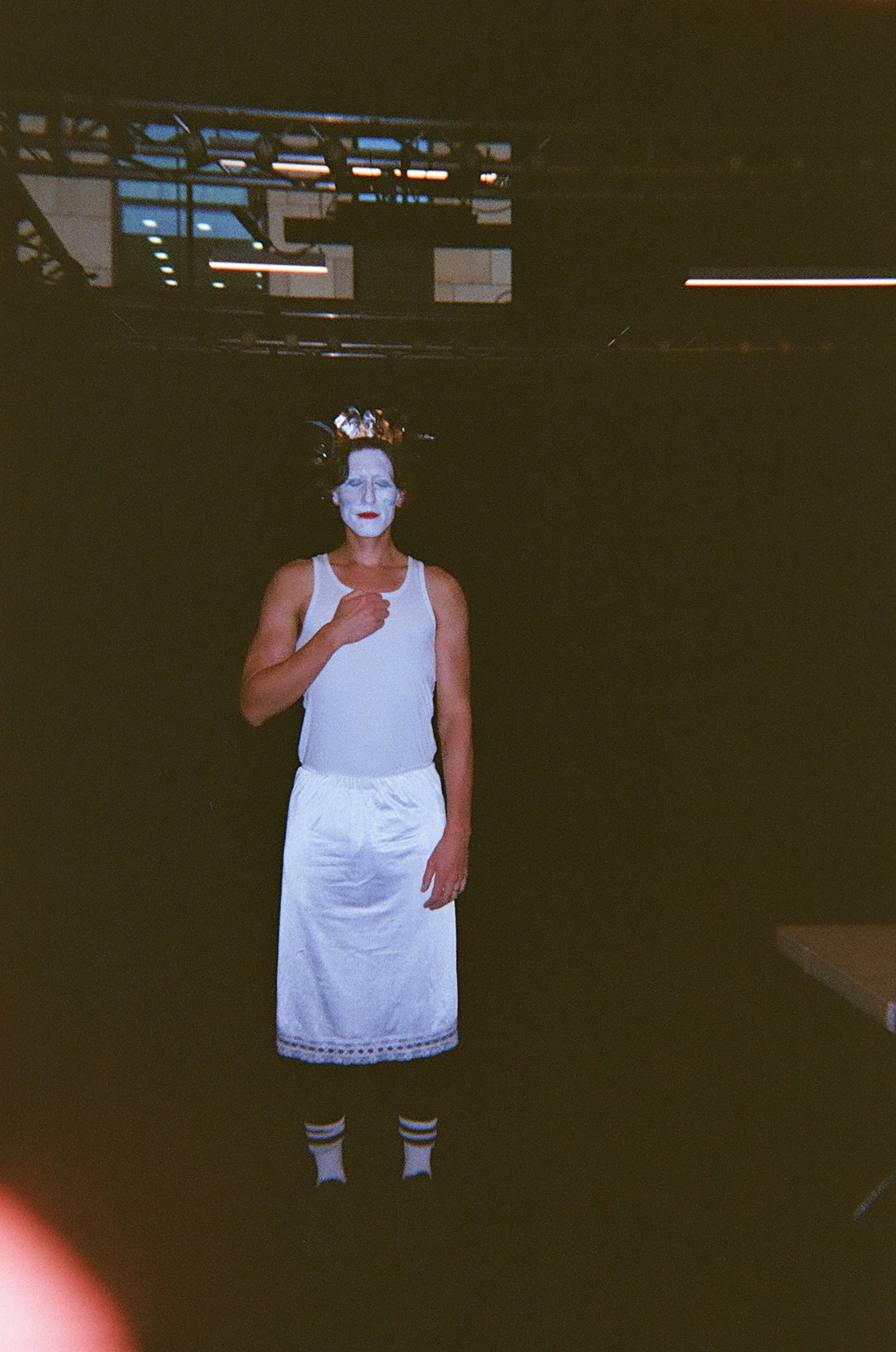 Person standing indoors against a dark background wearing white face paint, a gold crown, a sleeveless white top, white skirt with lace trim, striped socks, and dark shoes, with one hand touching their chest.