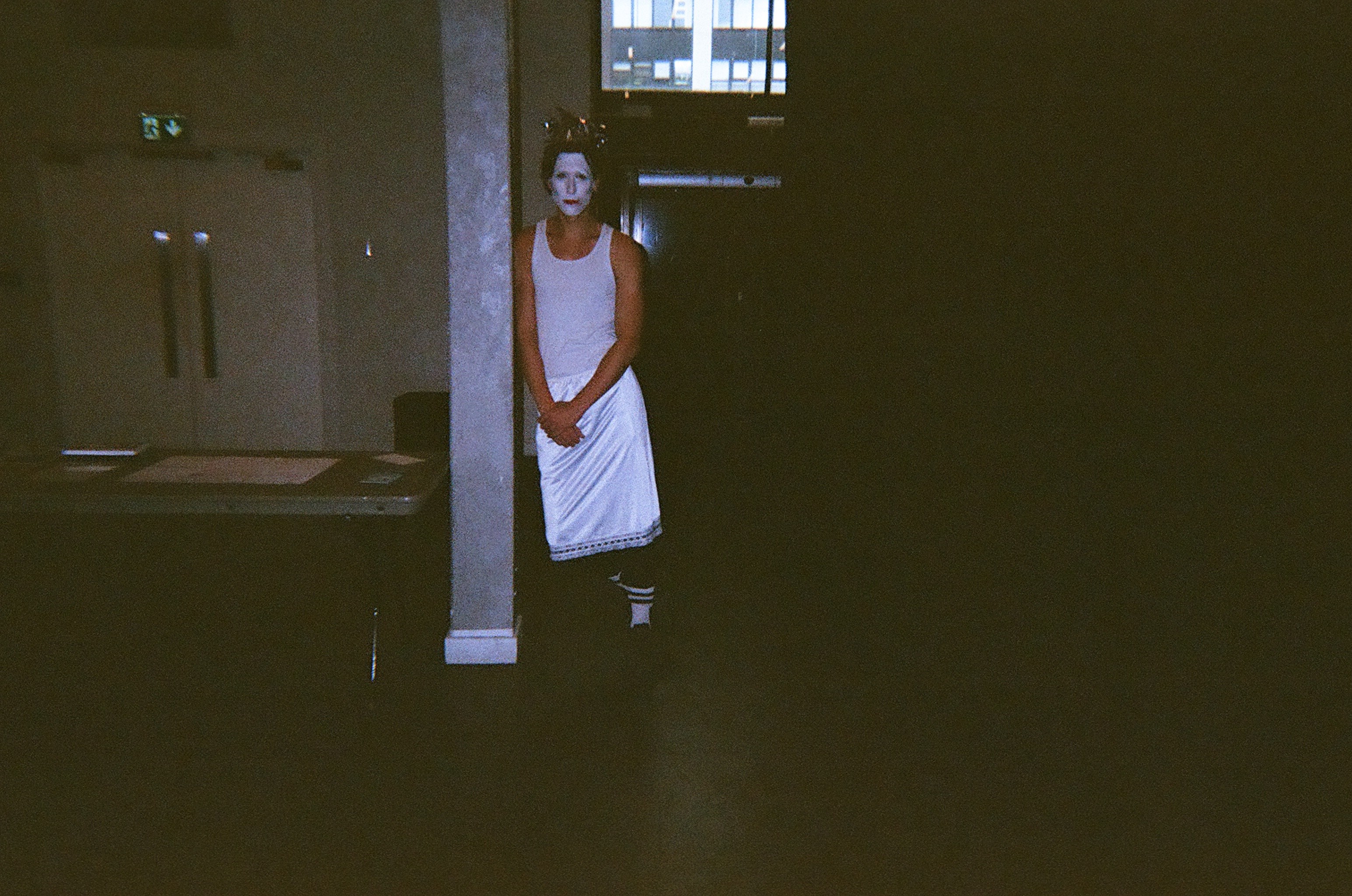 A person wearing a white sleeveless top and a white skirt with black and white striped socks, standing in a dimly lit room near a pillar, with their face painted white and a serious expression.
