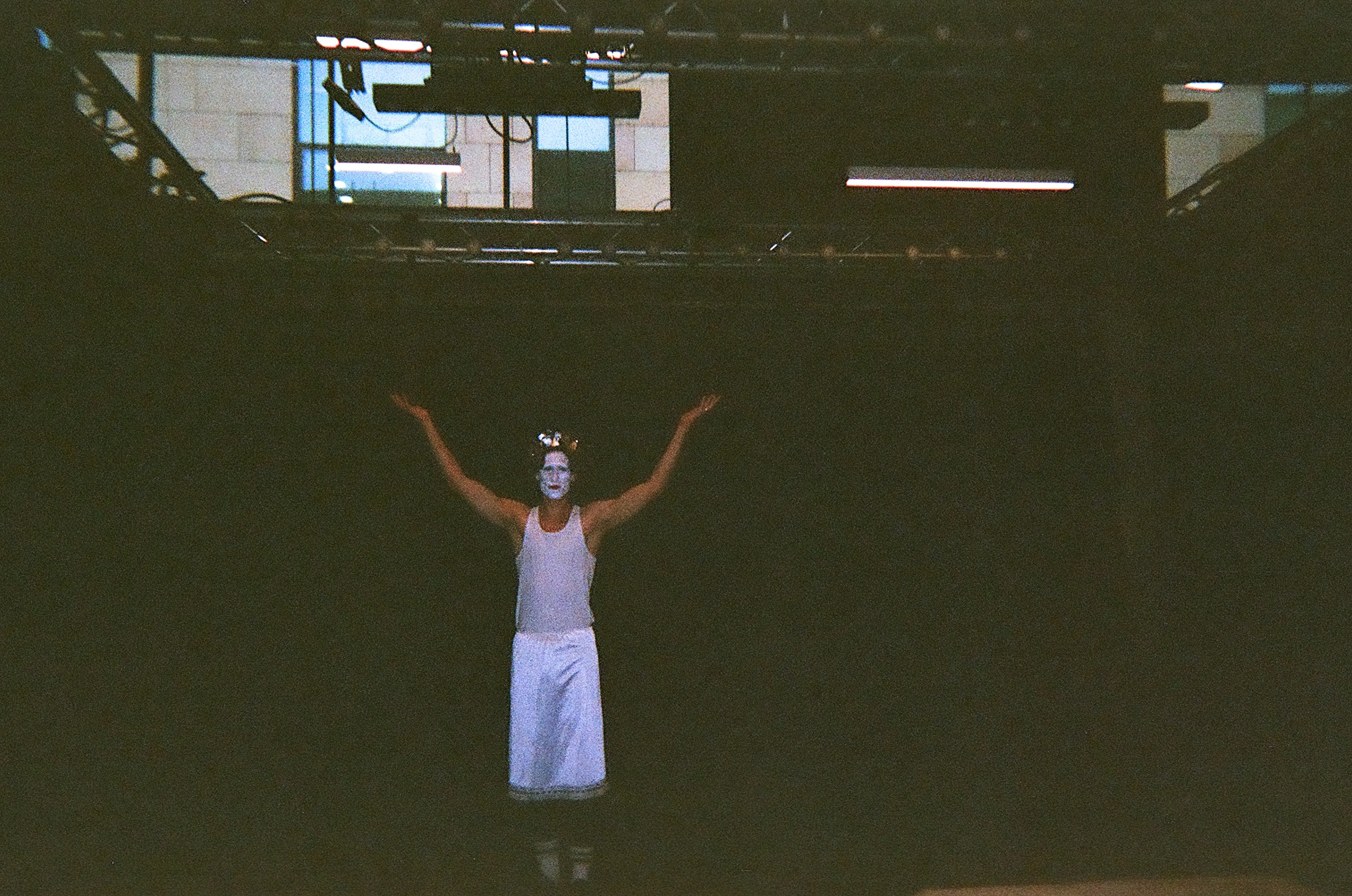 Person on stage with white face makeup and horns, wearing a white sleeveless top and skirt, standing with arms raised against a dark background.