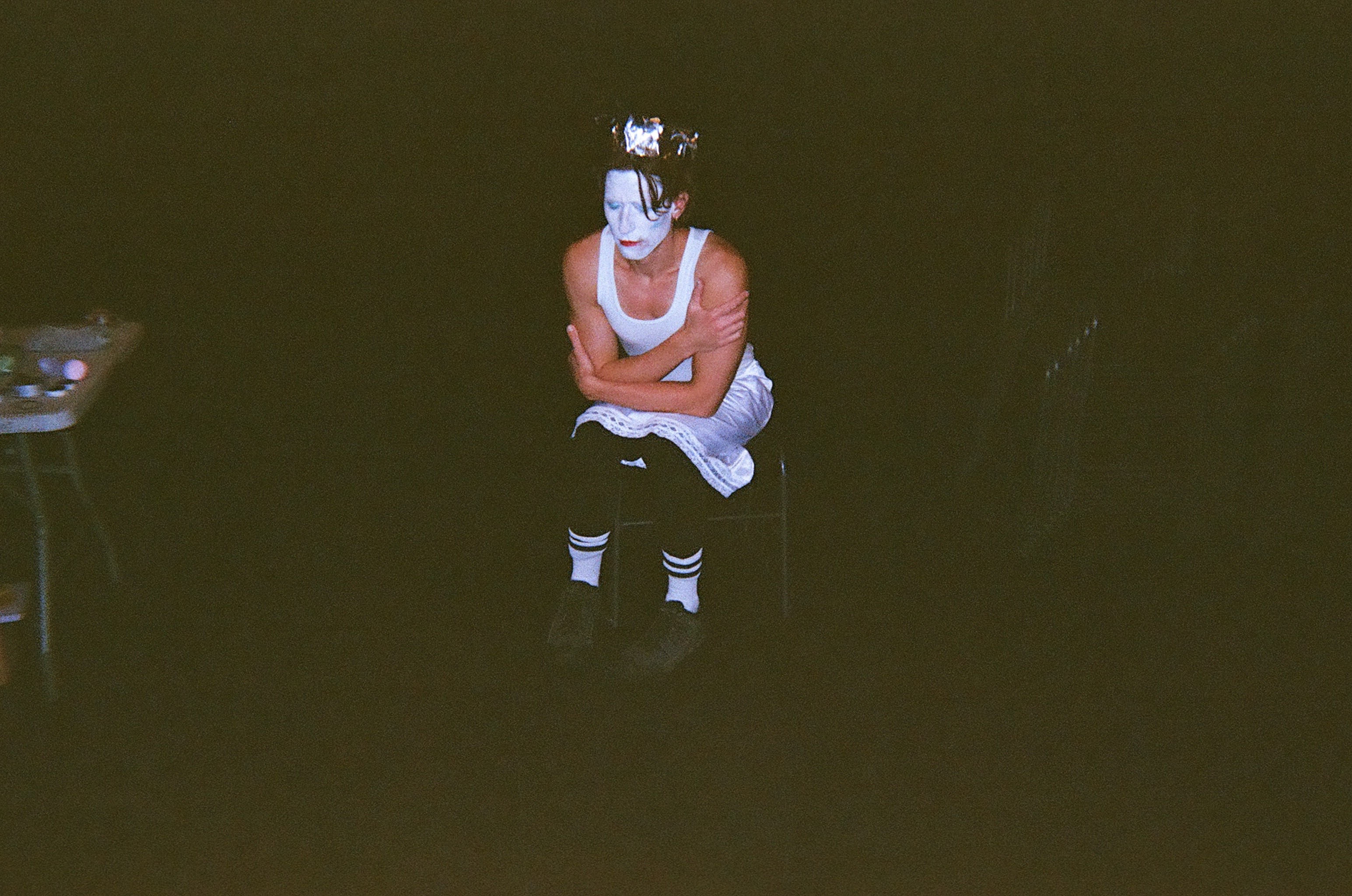 Person with white face paint sitting on a chair in a dark room, wearing a white tank top, black pants, white socks with black stripes, and a white apron, arms crossed over their chest with a serious expression.