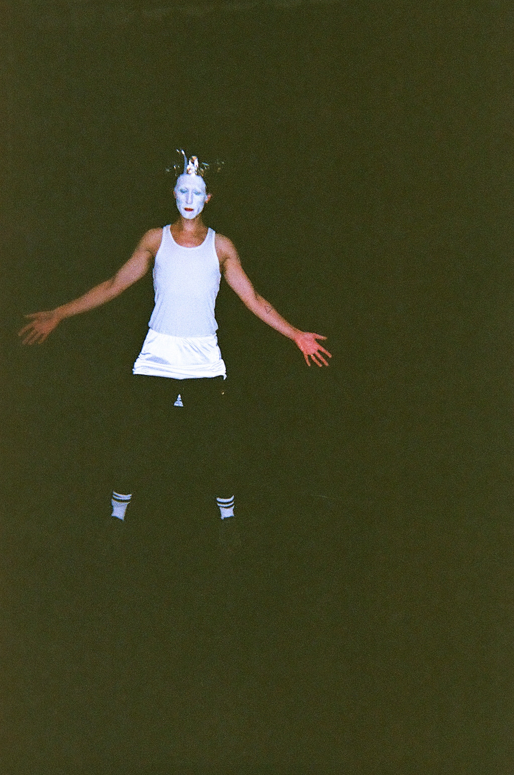 Person with white face paint and a headpiece with small horns or branches stands in darkness with arms outstretched, wearing a white tank top, white shorts, and black leggings with white striped socks.