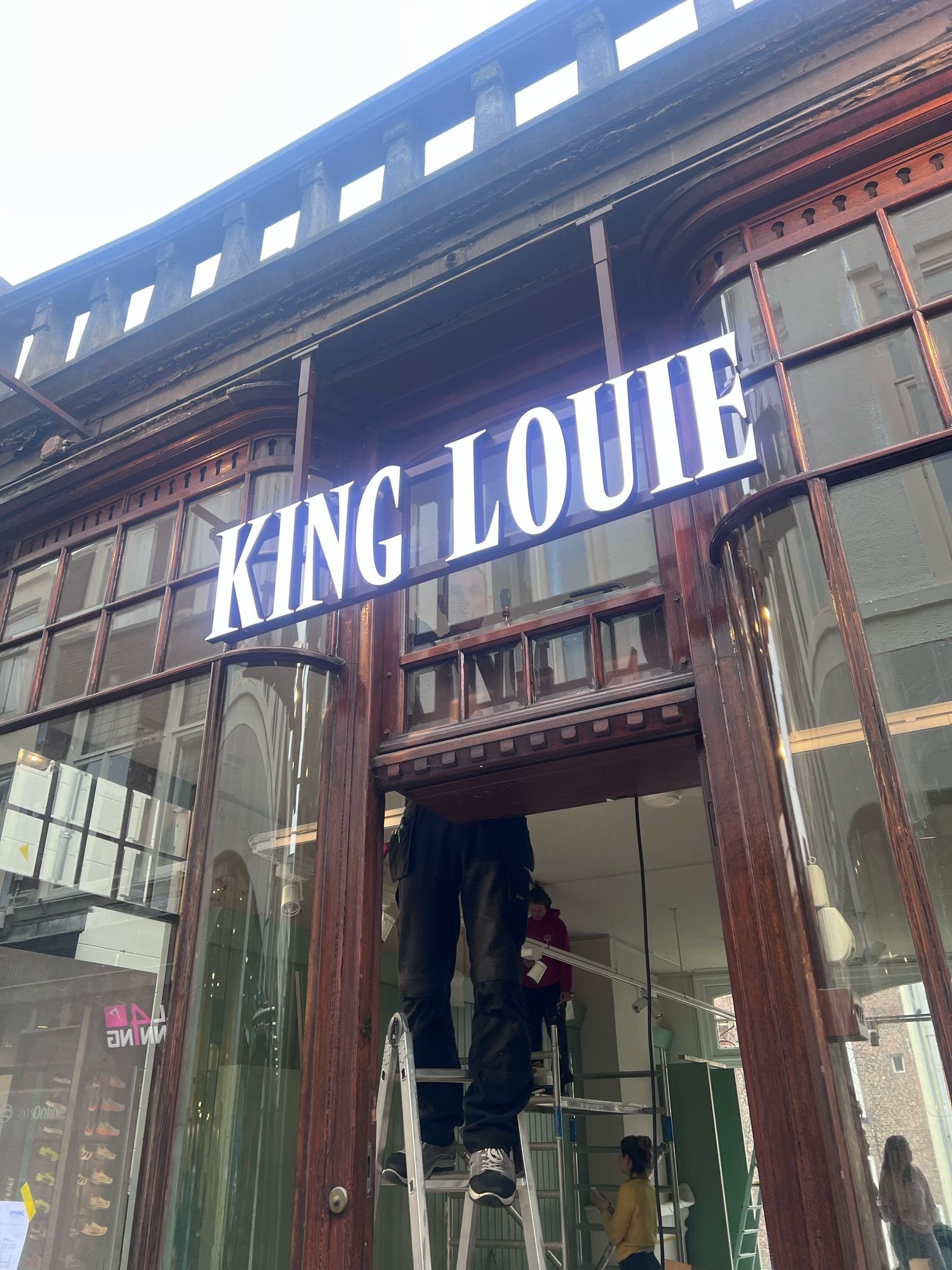 A person standing on a ladder inside a building under a large illuminated sign reading 'KING LOUIE' mounted on wooden framed windows and doors.