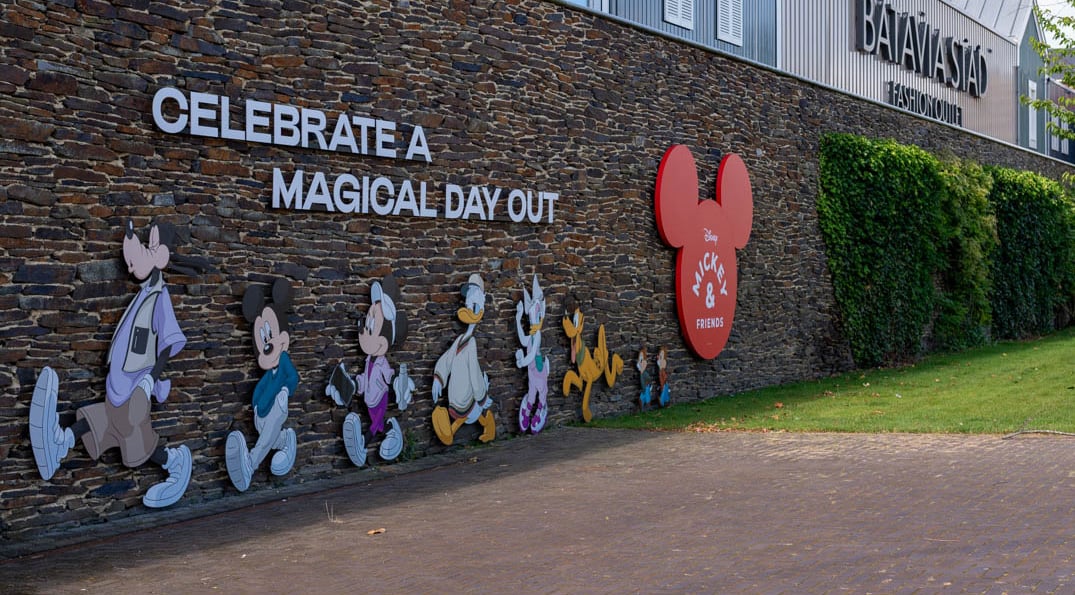 Stone wall featuring large colorful cutouts of Disney characters Goofy, Mickey Mouse, Minnie Mouse, Donald Duck, Daisy Duck, Pluto, and Chip and Dale walking in a line with the text 'Celebrate a magical day out' and a red Mickey Mouse head shape with 'Disney Mickey & Friends' near greenery, outside Batavia Stad Fashion Outlet.