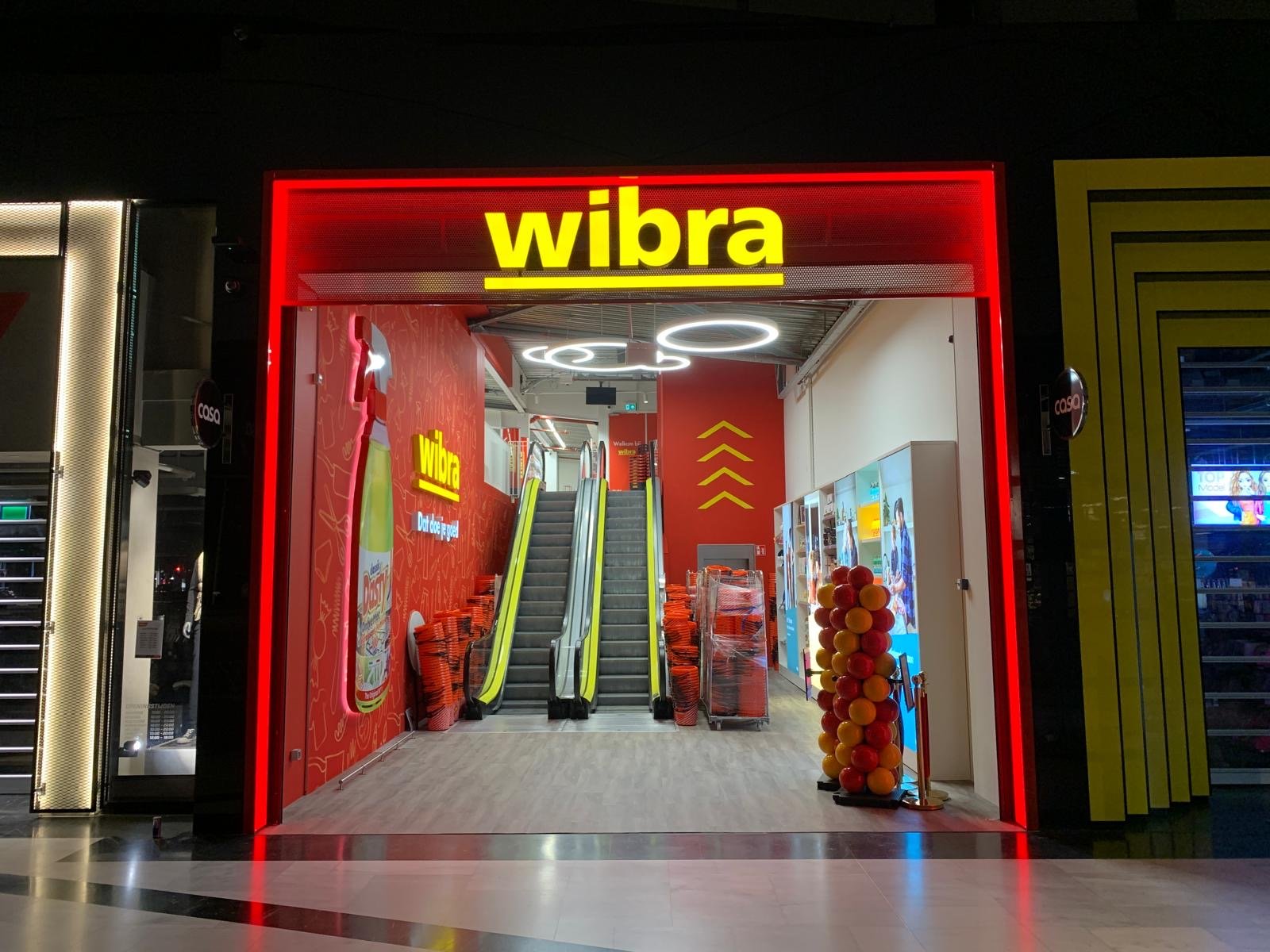 Entrance to a Wibra store with bright red and yellow signage, featuring escalators inside, stacks of orange shopping baskets, and a decorative column of red and yellow balloons on the right side.