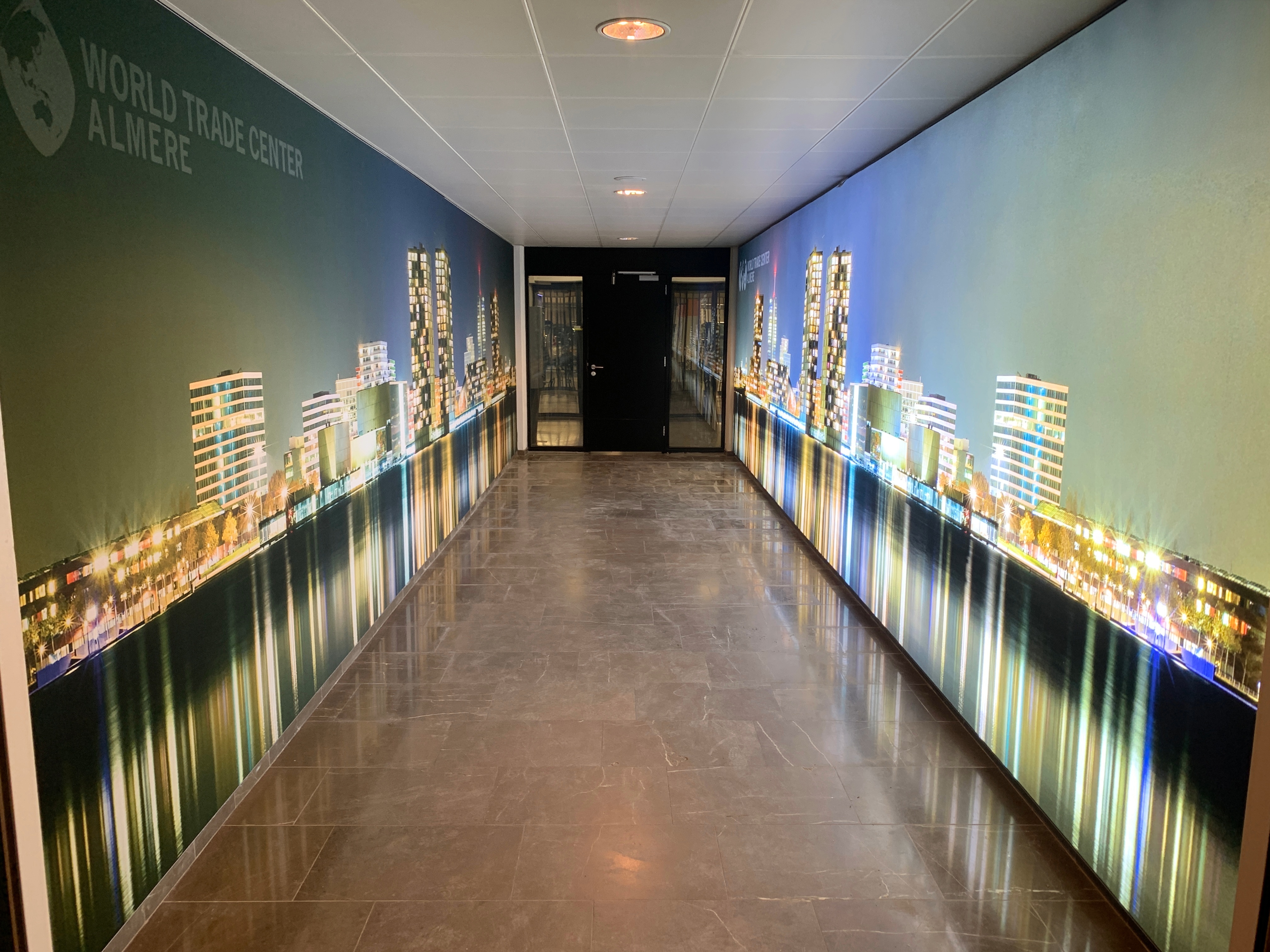 Long hallway with polished stone floor and ceiling lights, walls decorated with large photo murals of a city skyline at night reflecting on water, featuring tall buildings and colorful lights, with text 'World Trade Center Almere' on left wall.